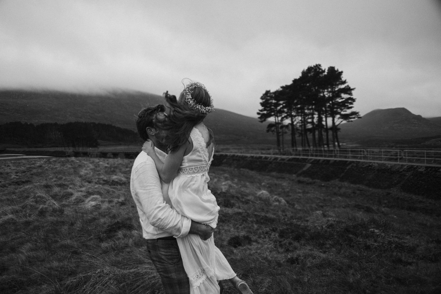 Black and white image of groom lifting bride outdoors near trees and misty mountains.