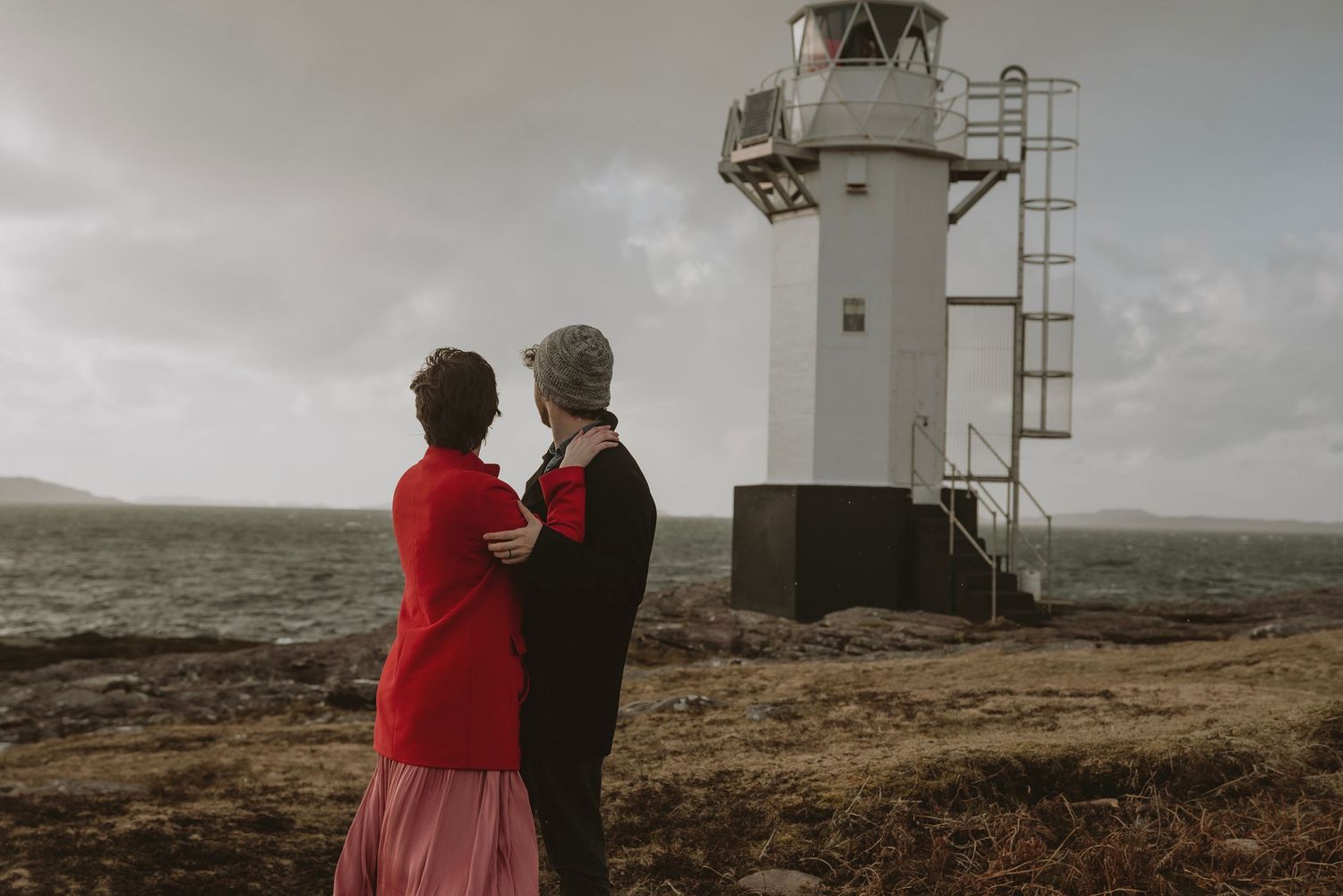 Couple embracing beside white lighthouse on rugged Scottish coastal shoreline.