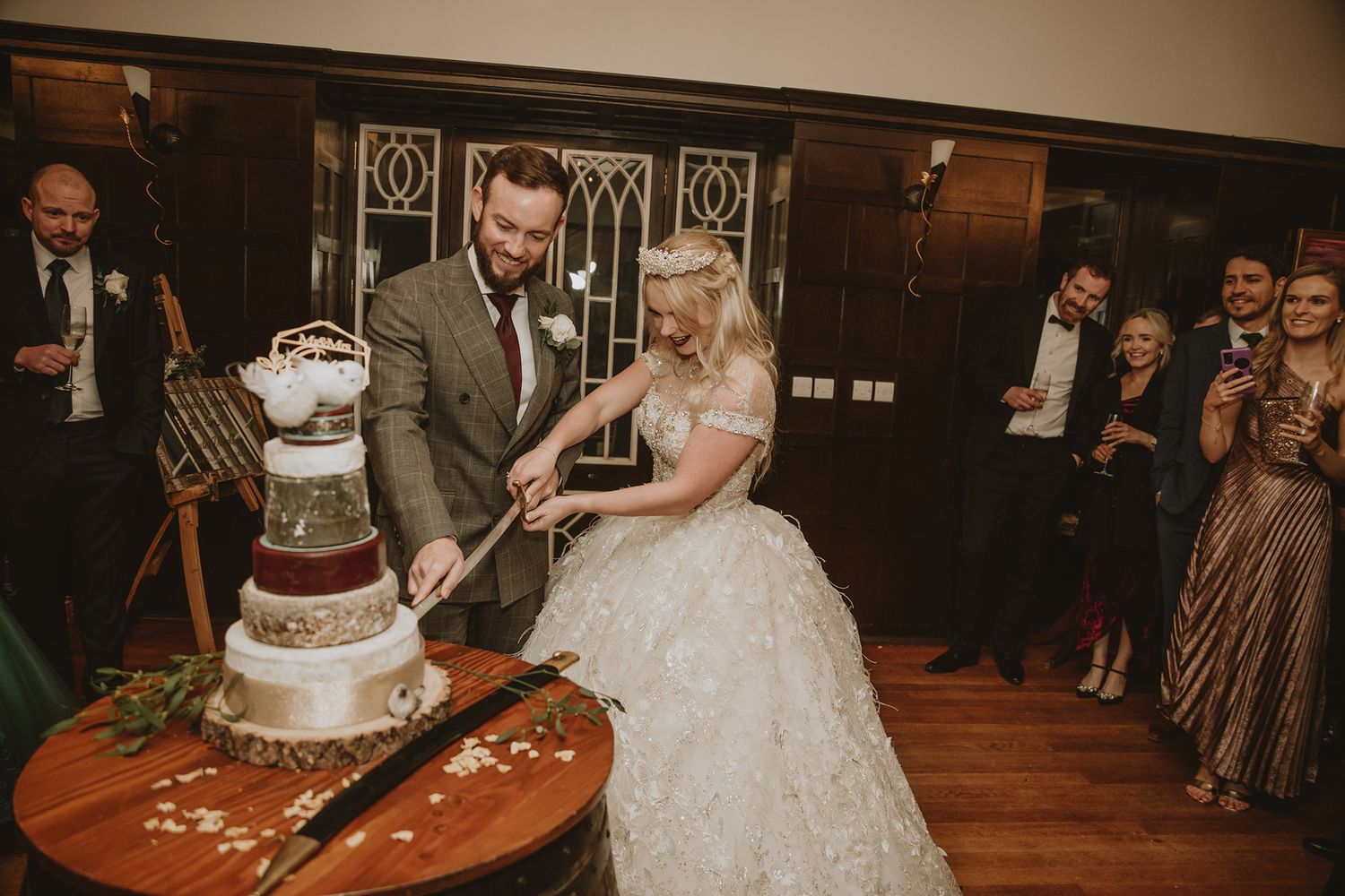 Bride and groom cutting their elegant multi-tiered wedding cake together while guests watch and celebrate in a wood-paneled venue.