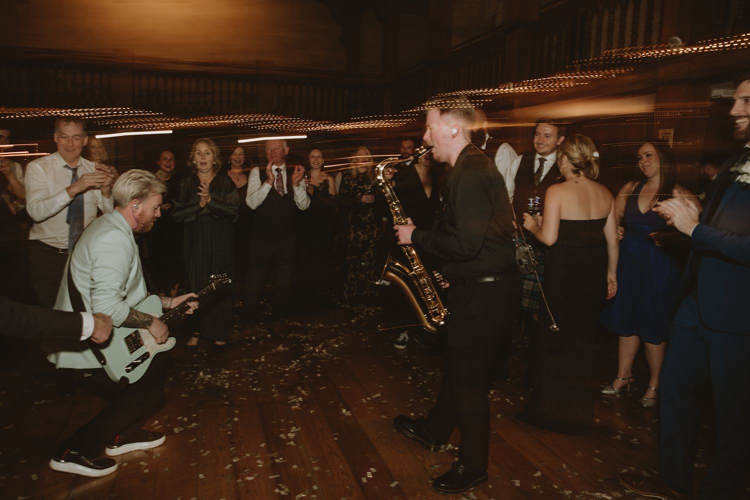 Live band performing with saxophonist and guitarist on wedding reception dance floor surrounded by cheering guests.