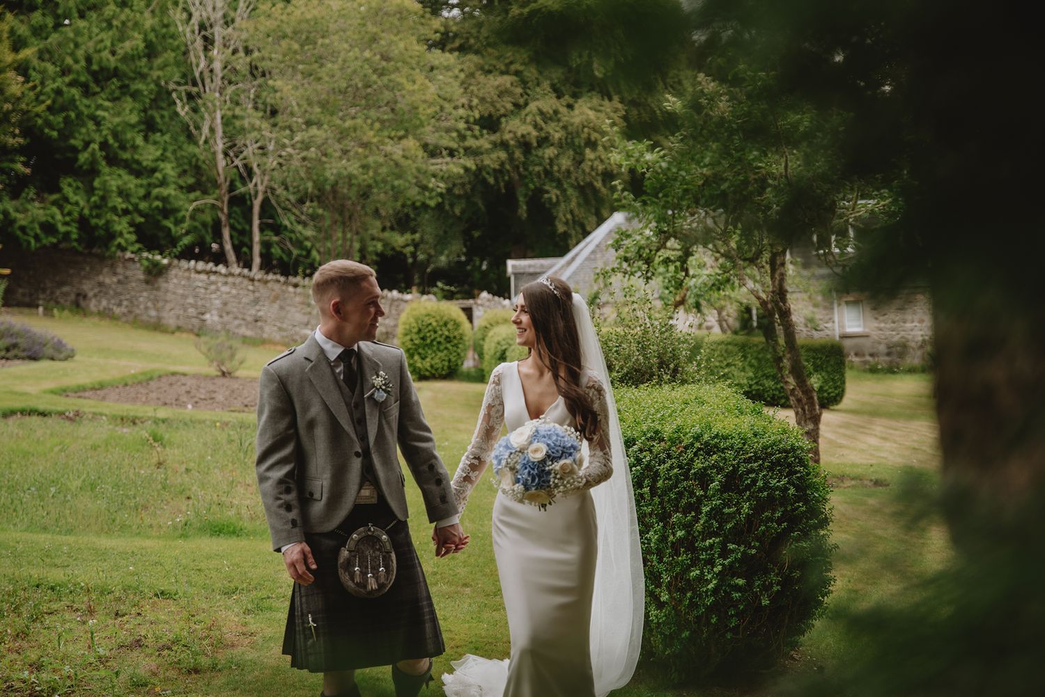 Bride and groom holding hands in lush garden, groom wearing traditional Scottish kilt, bride in white gown holding blue bouquet.