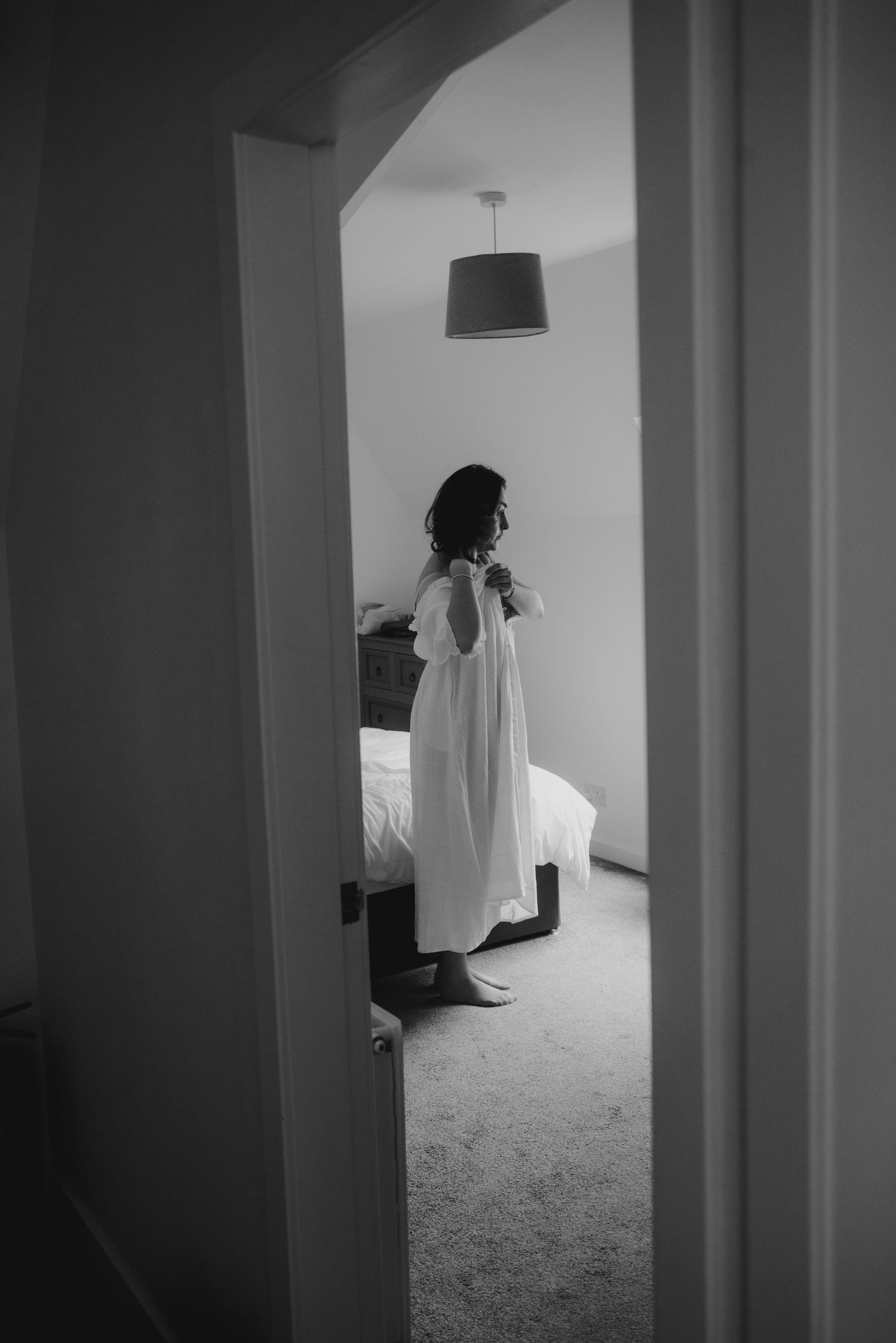 Black and white image of bride in white dress viewed through arched doorway in a bedroom.