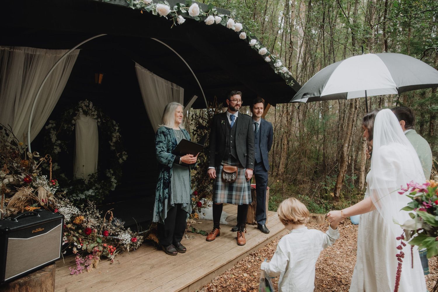 Groom in tartan kilt stands at woodland altar with officiant as bride approaches with child.
