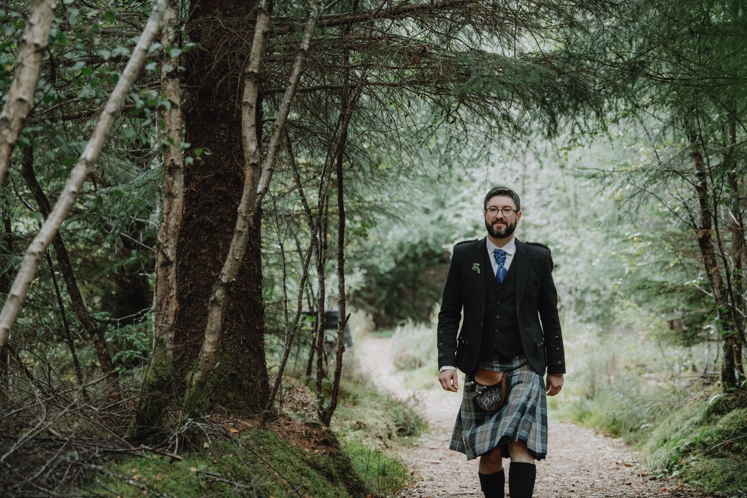 Bearded groom wearing a tartan kilt and dark jacket strolling down a woodland forest path.