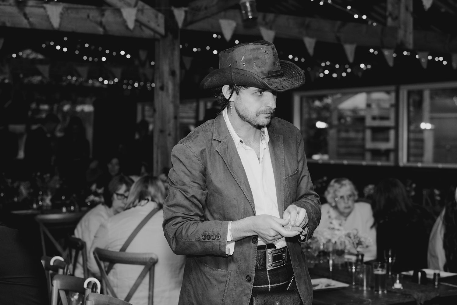 Man in cowboy hat and suit adjusting cufflinks at rustic wedding reception with fairy lights.