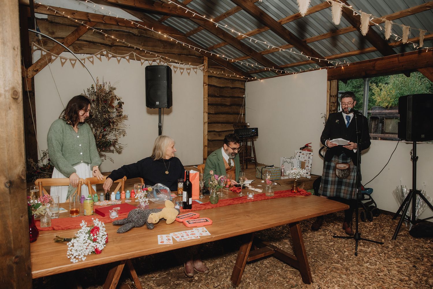 Wedding party seated at long rustic table as kilted speaker addresses guests in barn venue.