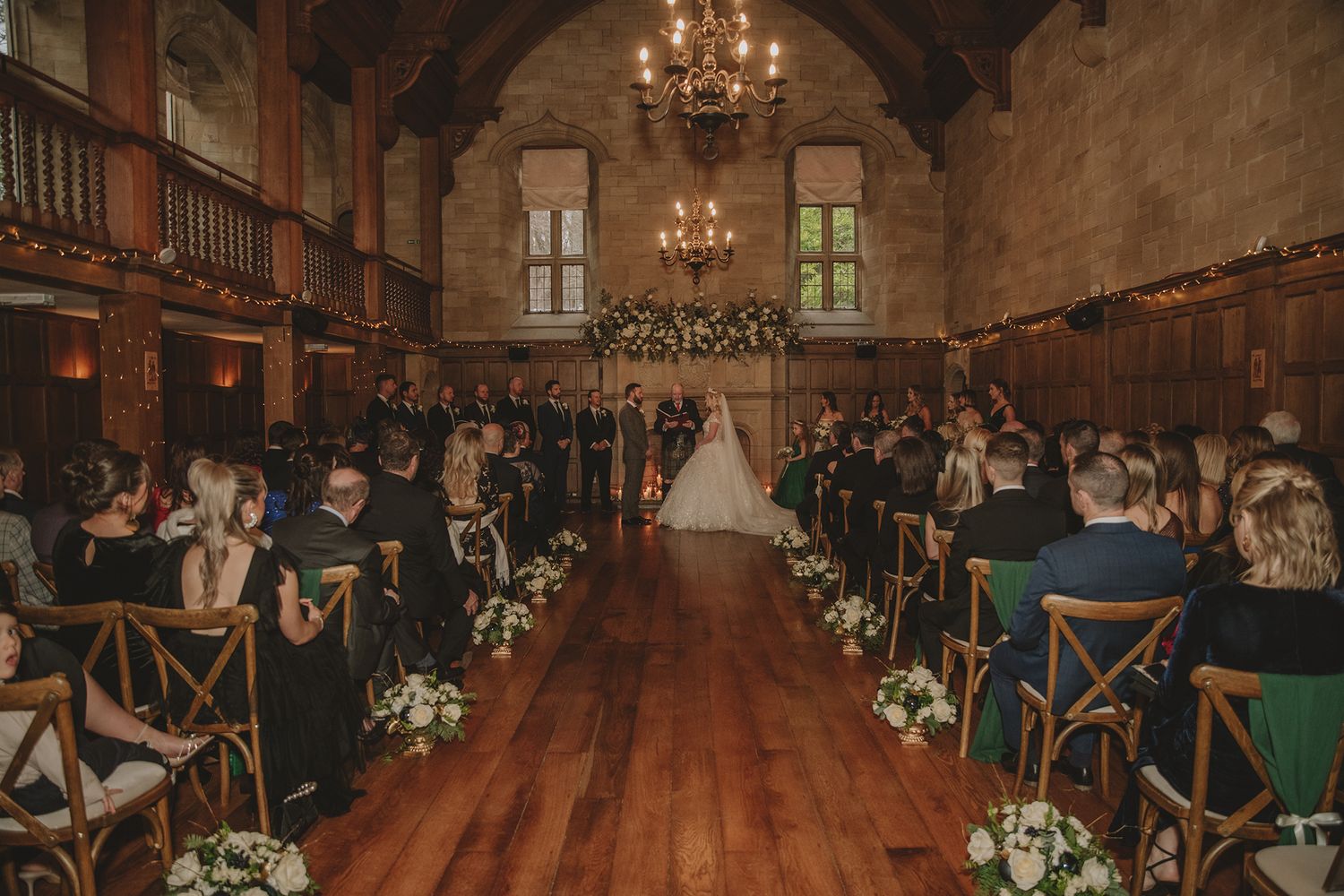 Elegant wedding ceremony in a grand hall with wooden floors, chandeliers, floral arch, and guests seated along the aisle.