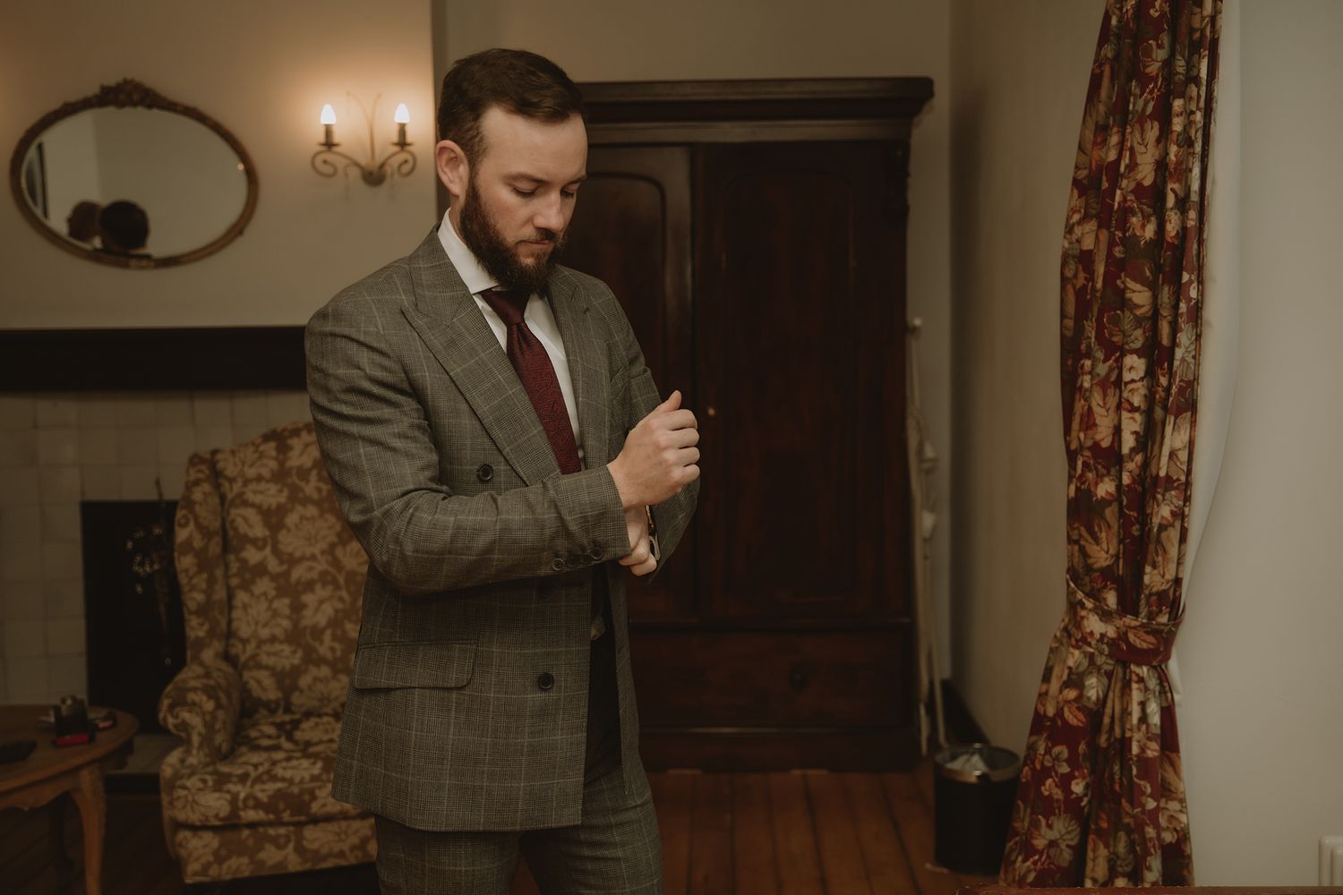 Groom in tweed suit adjusting cufflinks in elegant vintage room before wedding.