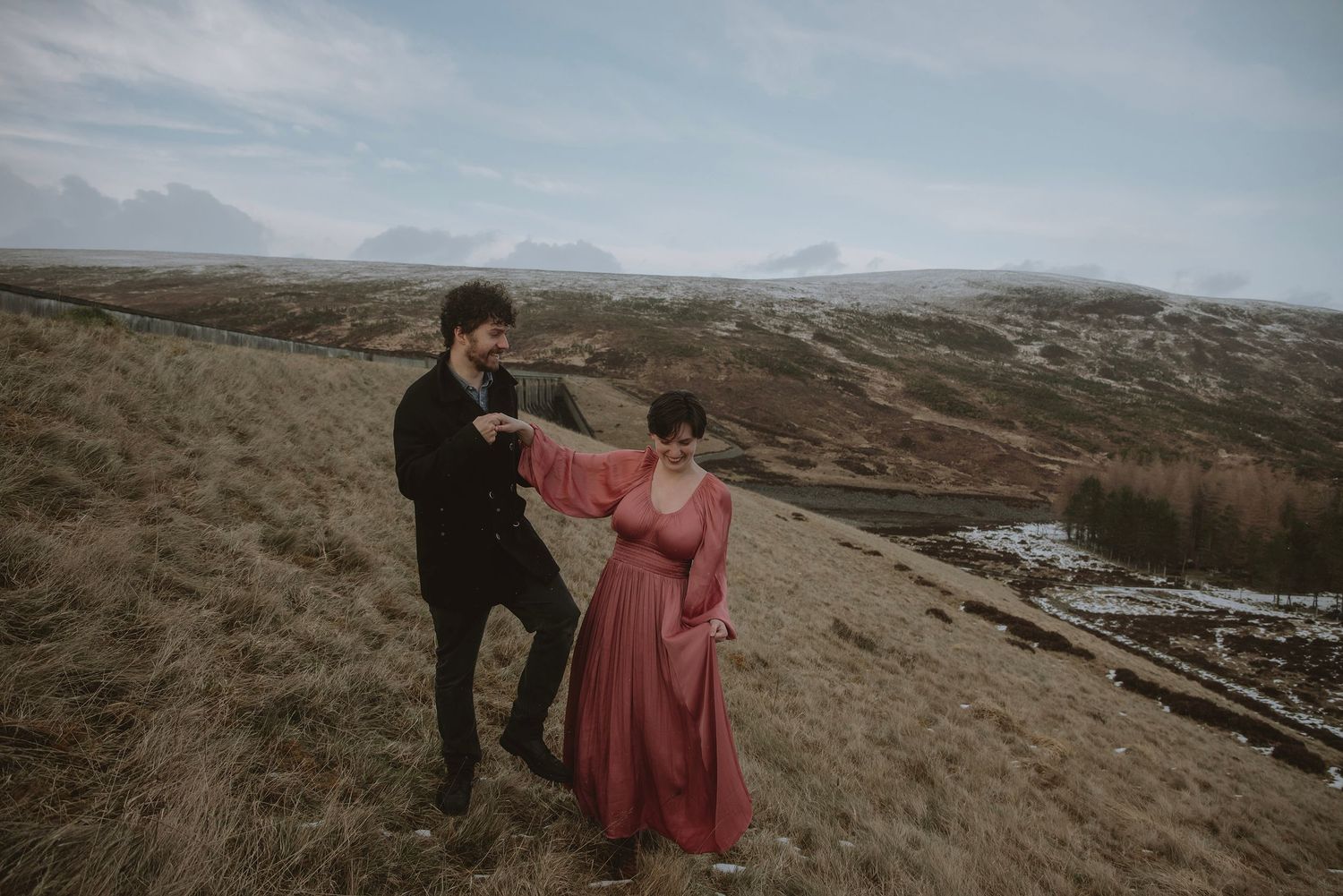 Couple dancing on a scenic hillside in Iceland, woman in red dress, man in black suit, moody landscape backdrop.