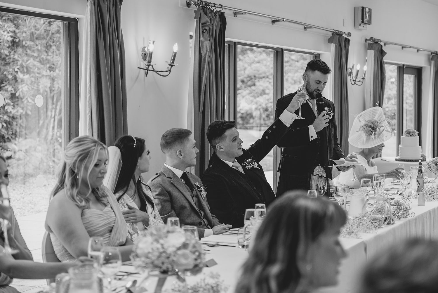 A man delivers a wedding speech to seated guests in an elegant venue, captured in black and white photography.
