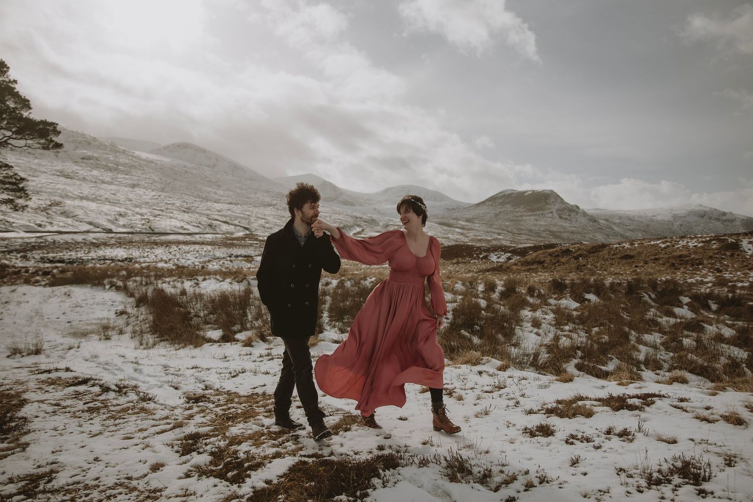 Couple dancing in snowy mountain landscape, woman in red dress, man in black coat, winter elopement photo.