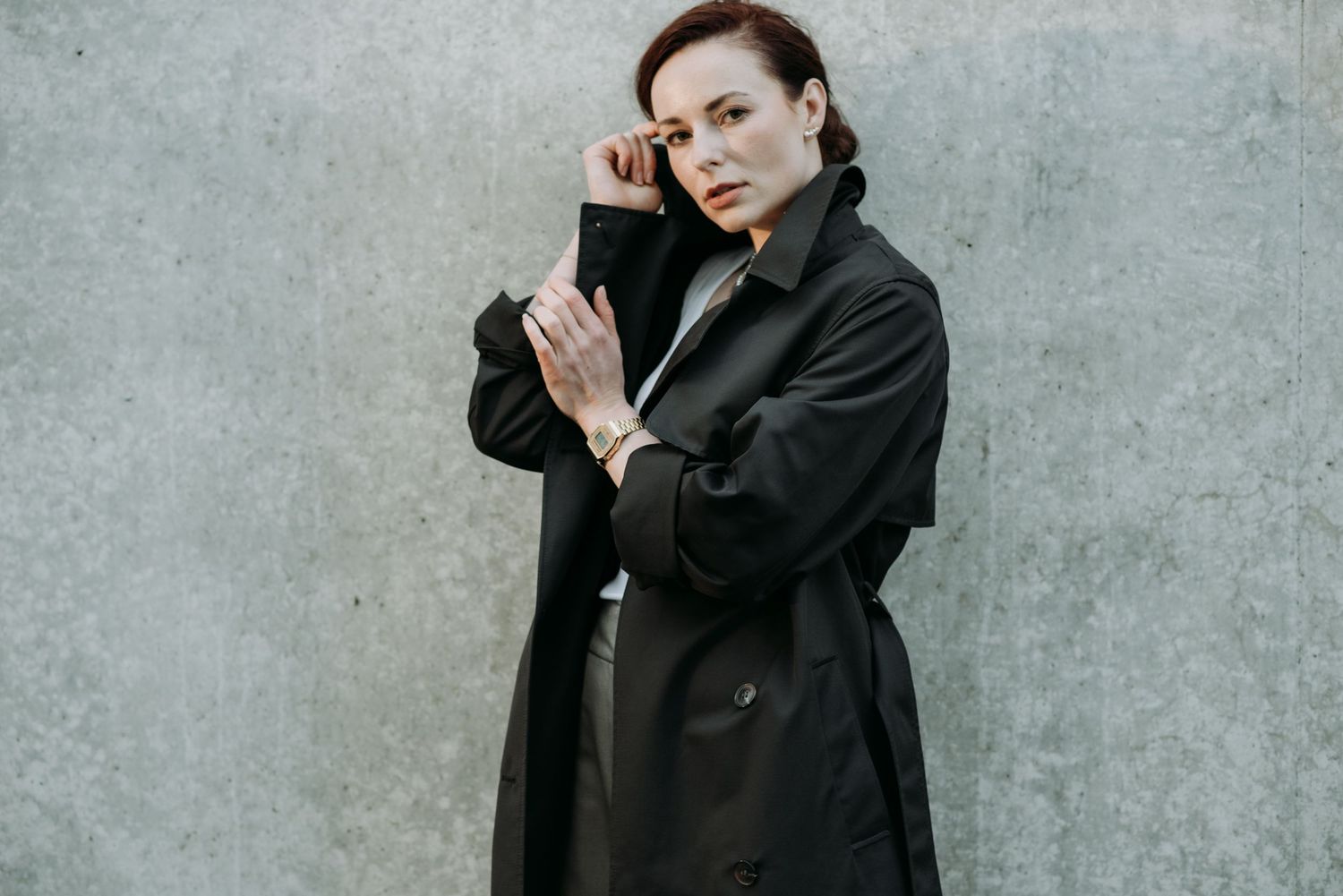 Stylish woman in black trench coat adjusts glasses against a grey concrete wall outdoors.