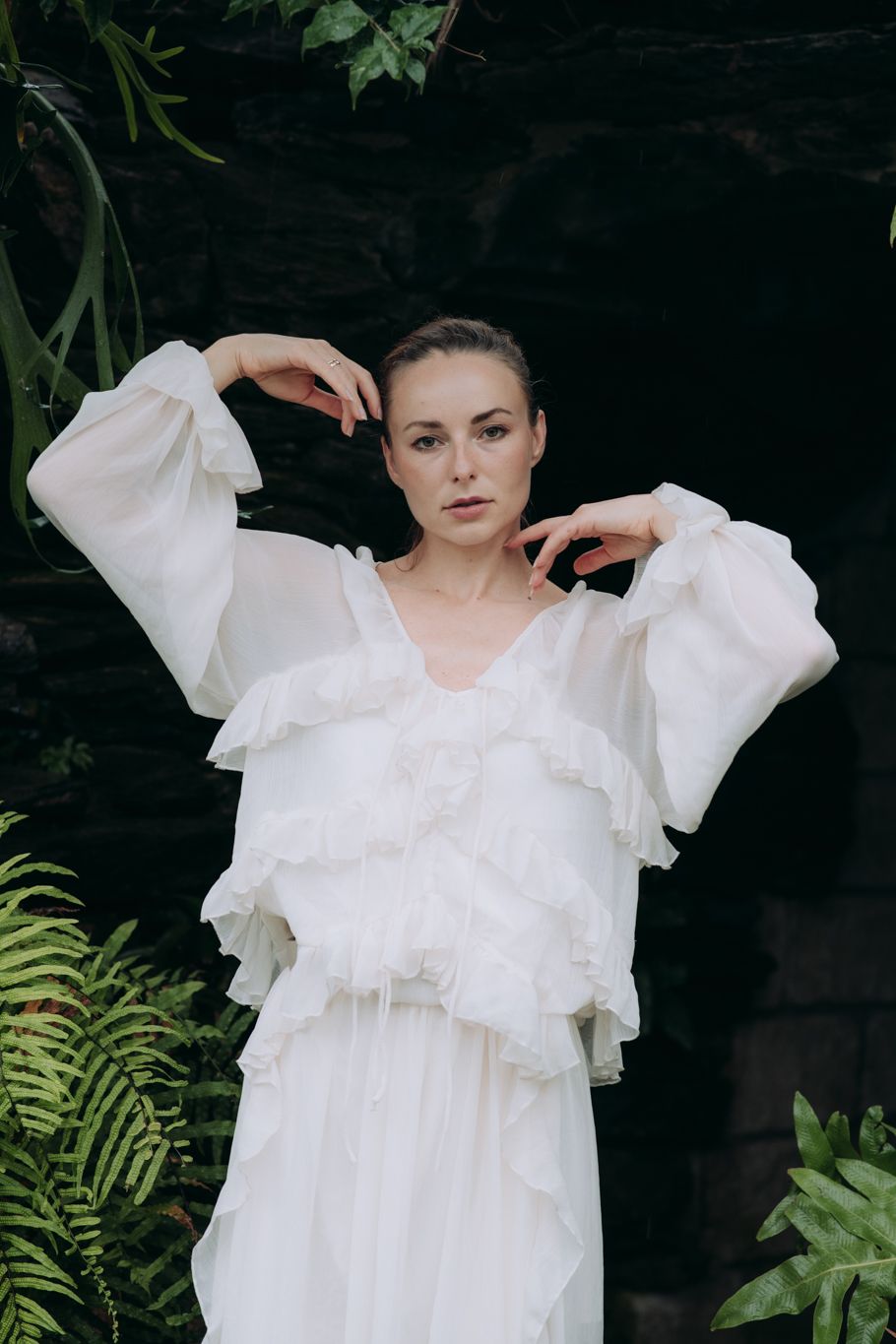 Woman in white ruffled balloon-sleeve dress posing with raised arms among dark green ferns and foliage.