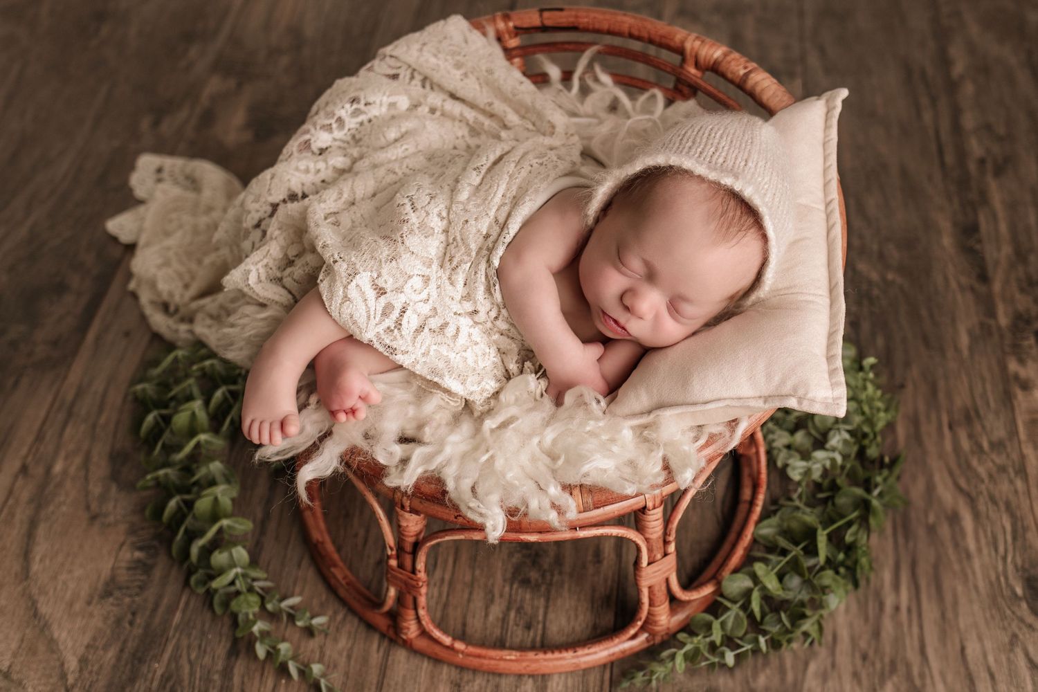 Baby girl curled up on bamboo papasan chair with soft fur and lace blankets. Wood floor background with eucalyptus garland.