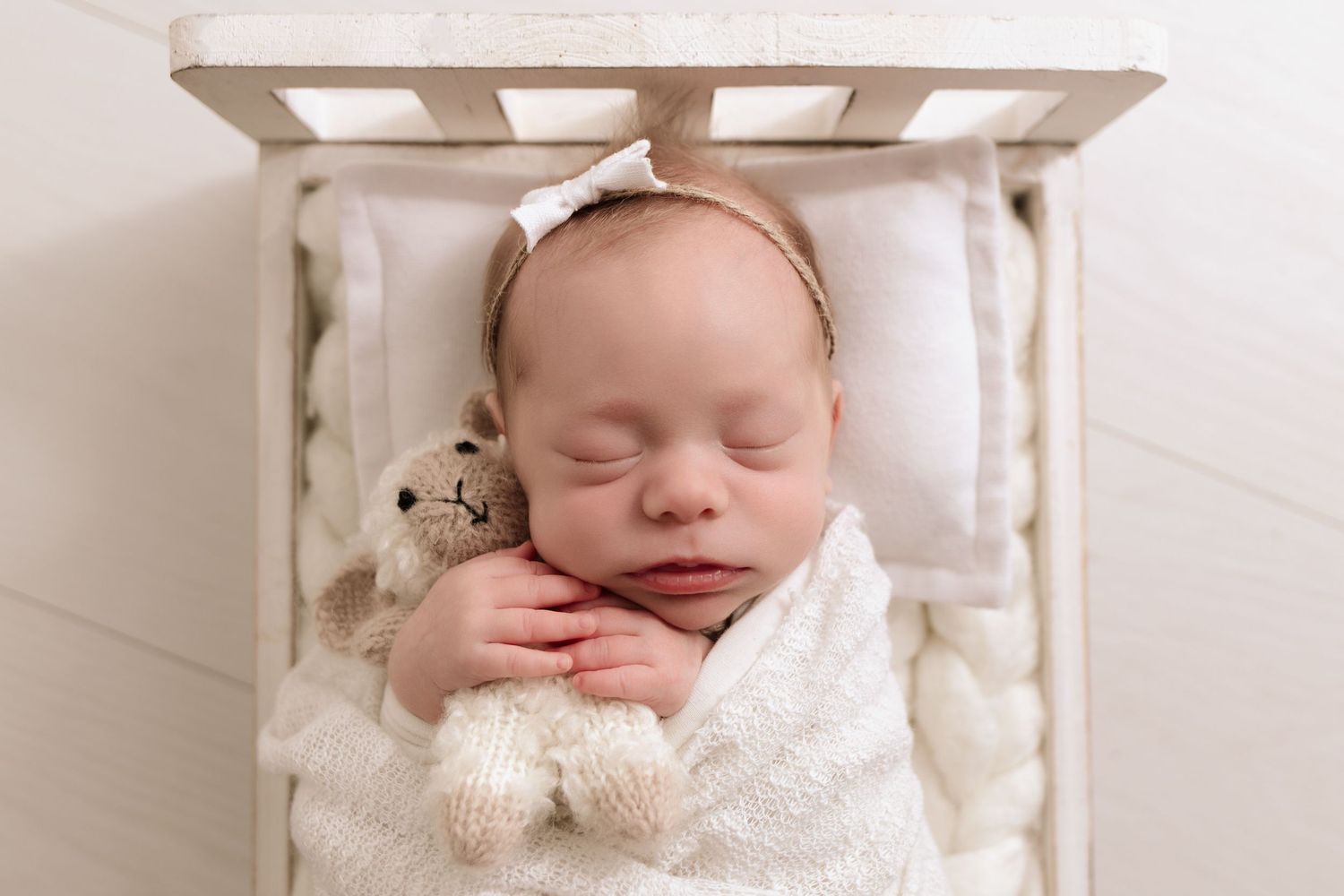Newborn girl photography session. Closeup of baby girl, swaddled in lace fabric, with her tiny hands wrapped around a tiny stuffed lamb.