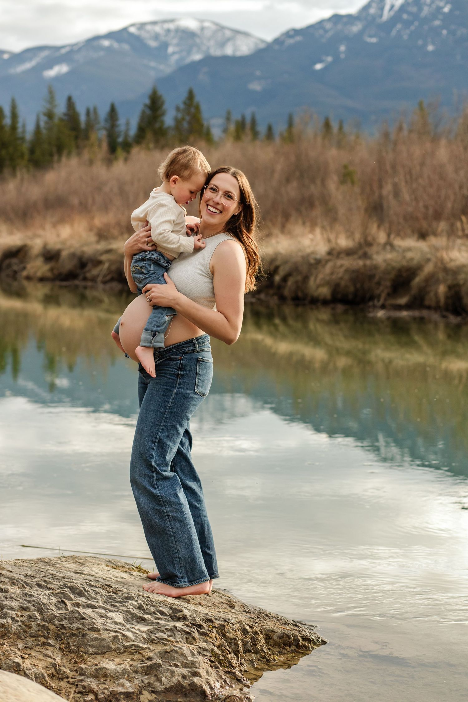 An expecting mom holding her toddler during a photo session with a beautiful mountain backdrop in Fairmont Hot Springs.