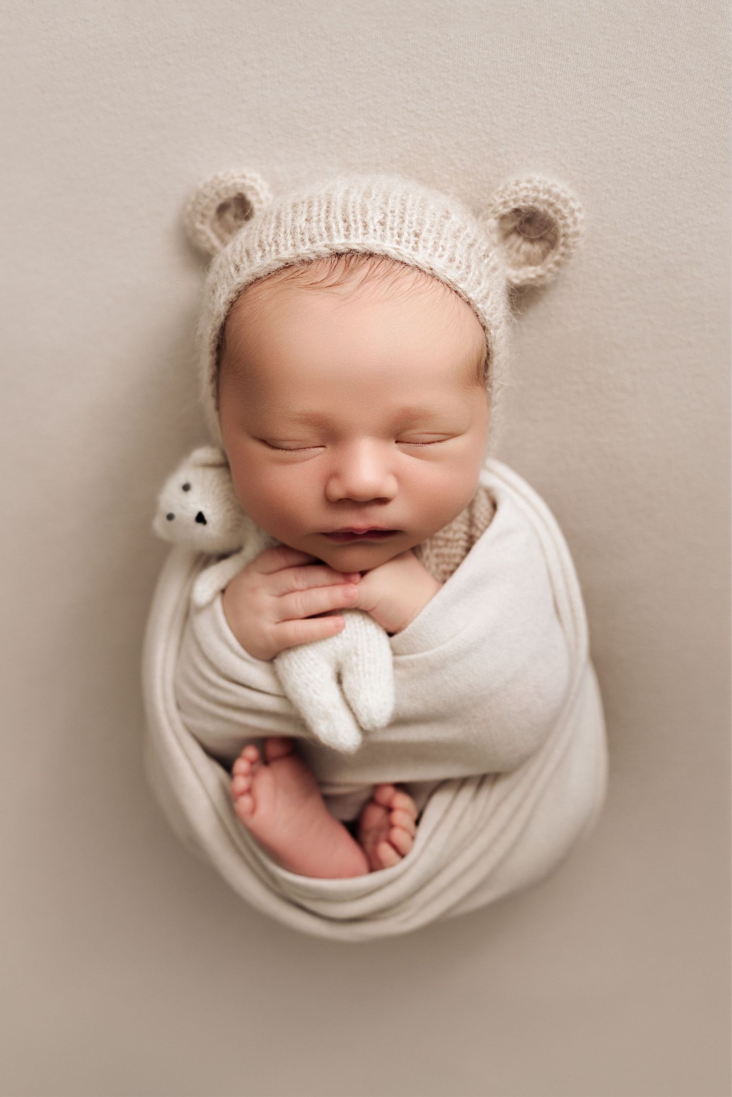 Newborn baby boy swaddled in neutral coloured fabric holding a tiny teddy bear. Baby boy is wearing a knit hat with teddy bear ears.