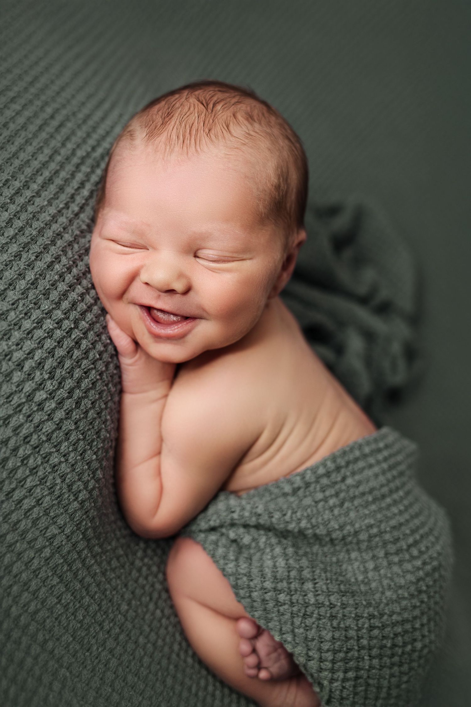 Newborn baby boy photo session. Sleeping baby boy is curled up on green fabric, hand resting under cheek and an adorable newborn smile.
