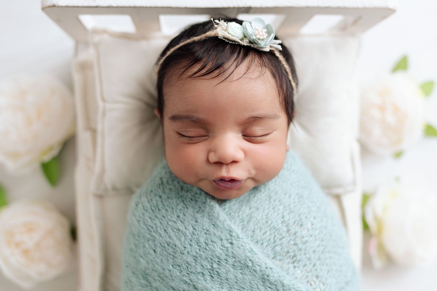 Gorgeous newborn baby girl with thick black hair, swaddled in a soft blue knit wrap. Baby girl has a cute expression with puckered lips.