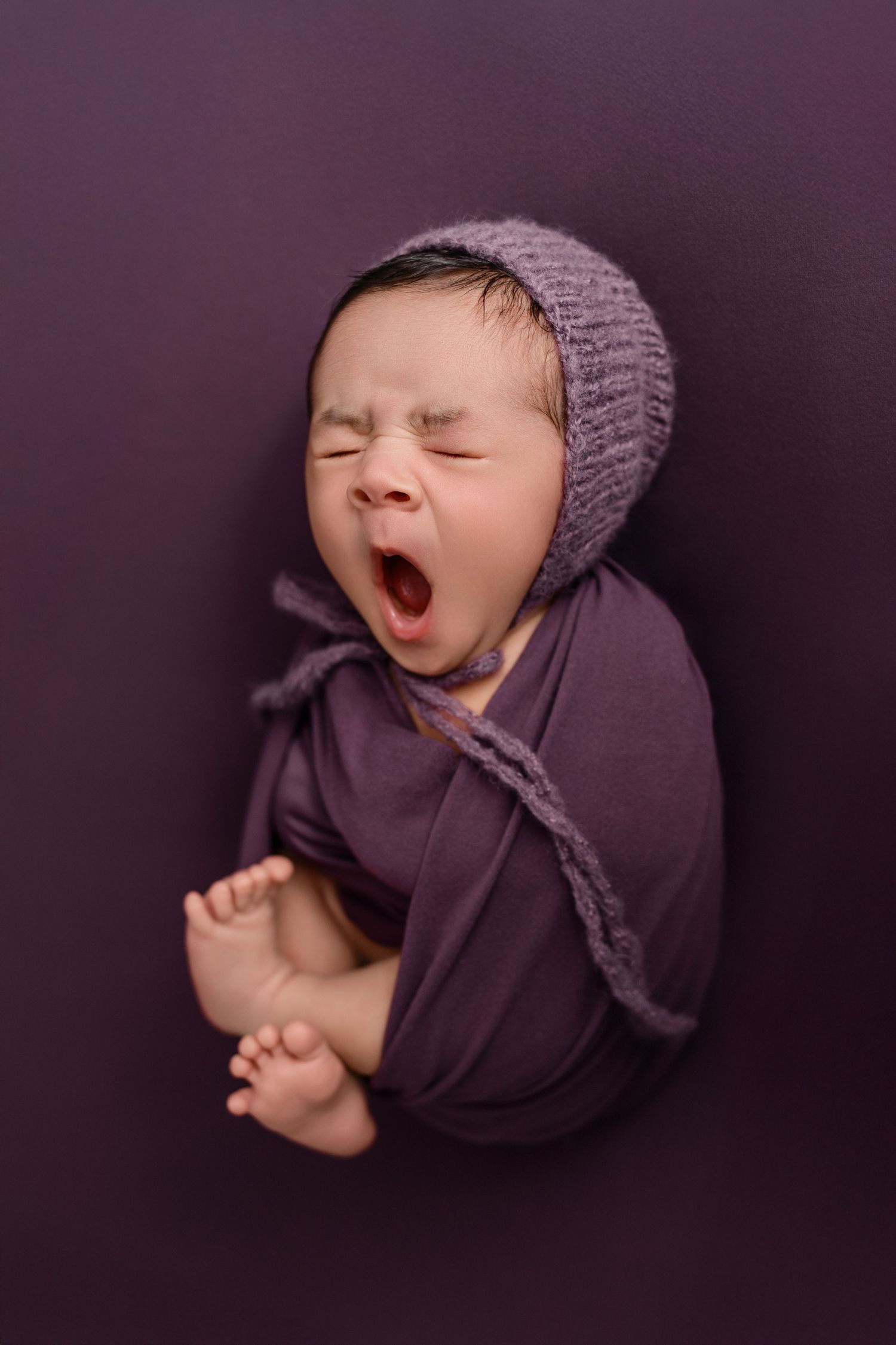 Yawning baby girl, swaddled in soft purple wrap with bonnet, during a posed newborn studio session at The Essence Photography by Mikelle.