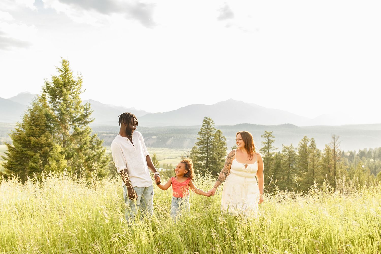 Family of 3, mom dad and young daughter smile at each other as they walk through a grassy field with trees and mountains in the background.