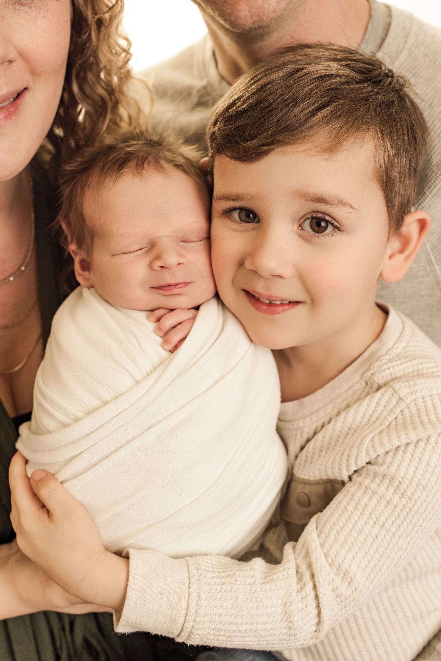 Zoomed in shot of big brother holding his newborn baby brother, swaddled in white, both boys are smiling. Parents are holding both boys.