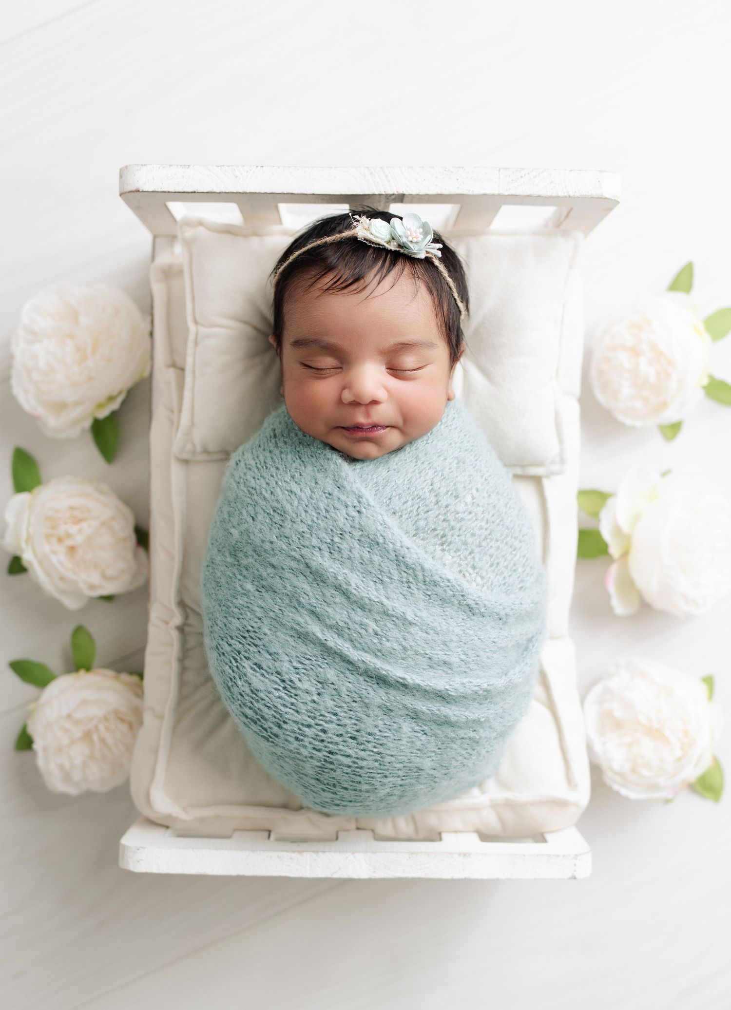 Newborn baby girl perfectly swaddled in soft knit blue wrap, on a tiny white bed, surrounded by fully opened white roses.