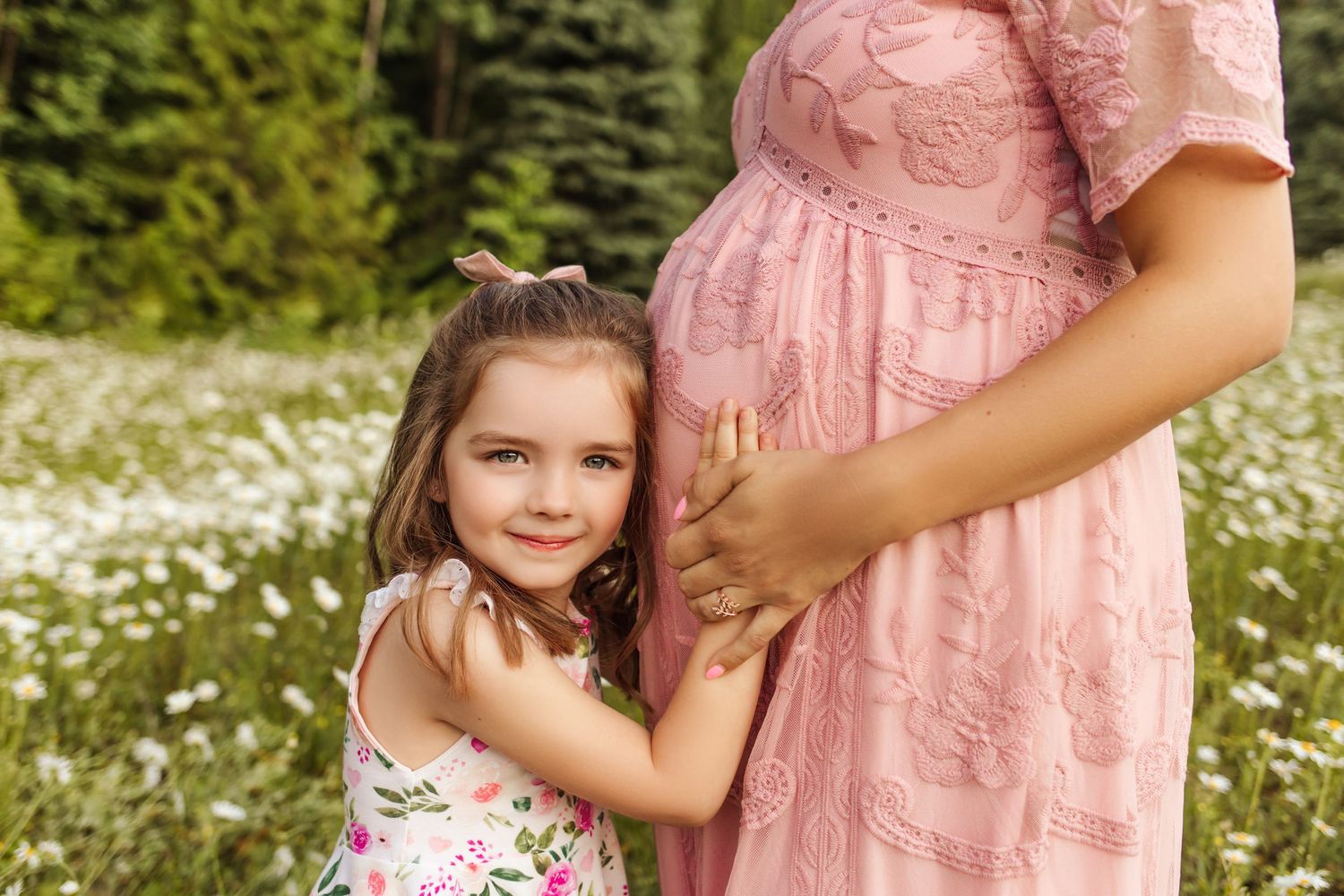 Beautiful blue-eyed girl hugs her mom's tummy. Mom is pregnant, wearing a pink maternity gown. Mom and daughter have hands on mom's tummy.