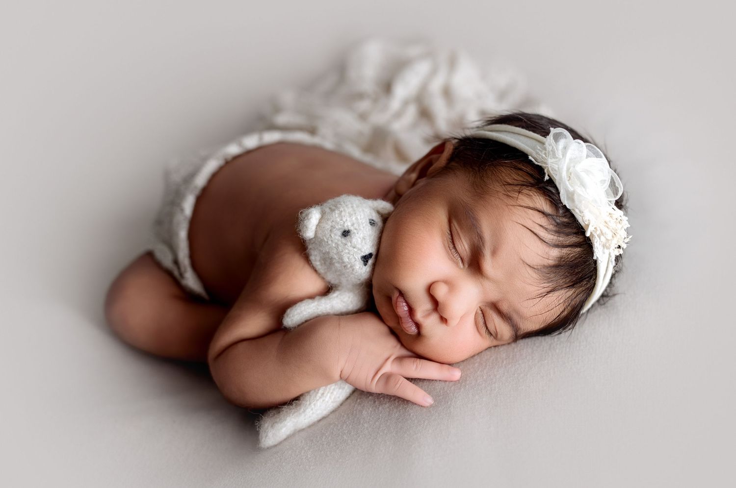 Beautiful dark skinned newborn baby girl in taco pose, snuggling a tiny knit teddy bear. Baby appears to have a slight scowl on her face.