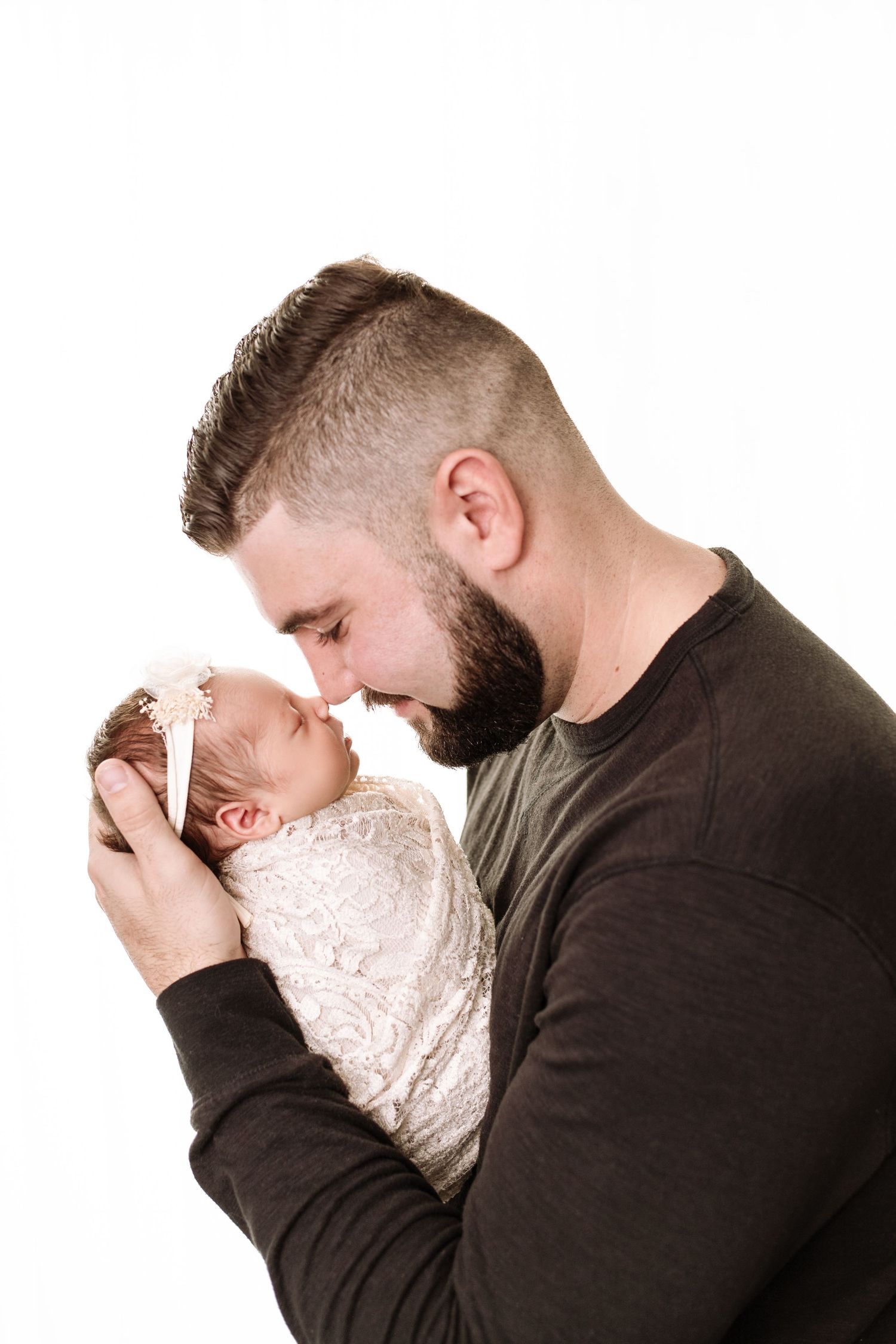 Dad holds newborn baby girl, swaddled with a pretty headband. Dad smiles, cradling baby's head in his hand and their noses touch.