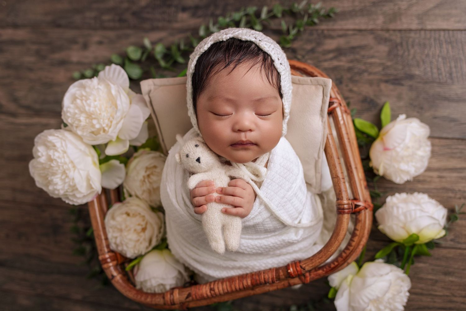 Gorgeous image of newborn wrapped and set in bamboo basket holding tiny knit teddy bear, sleeping peacefully, surrounded by flowers.