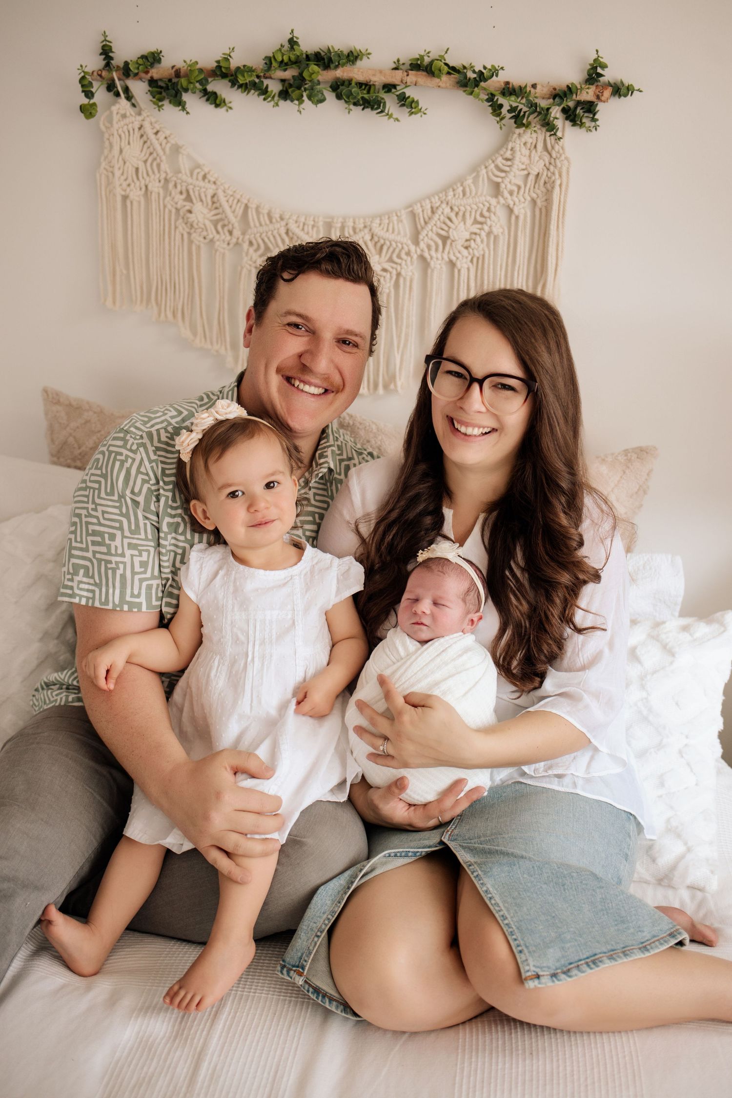 Family of four cuddled up on a bed for a newborn photo session. Newborn girl is swaddled in white and big sister is in a white dress..