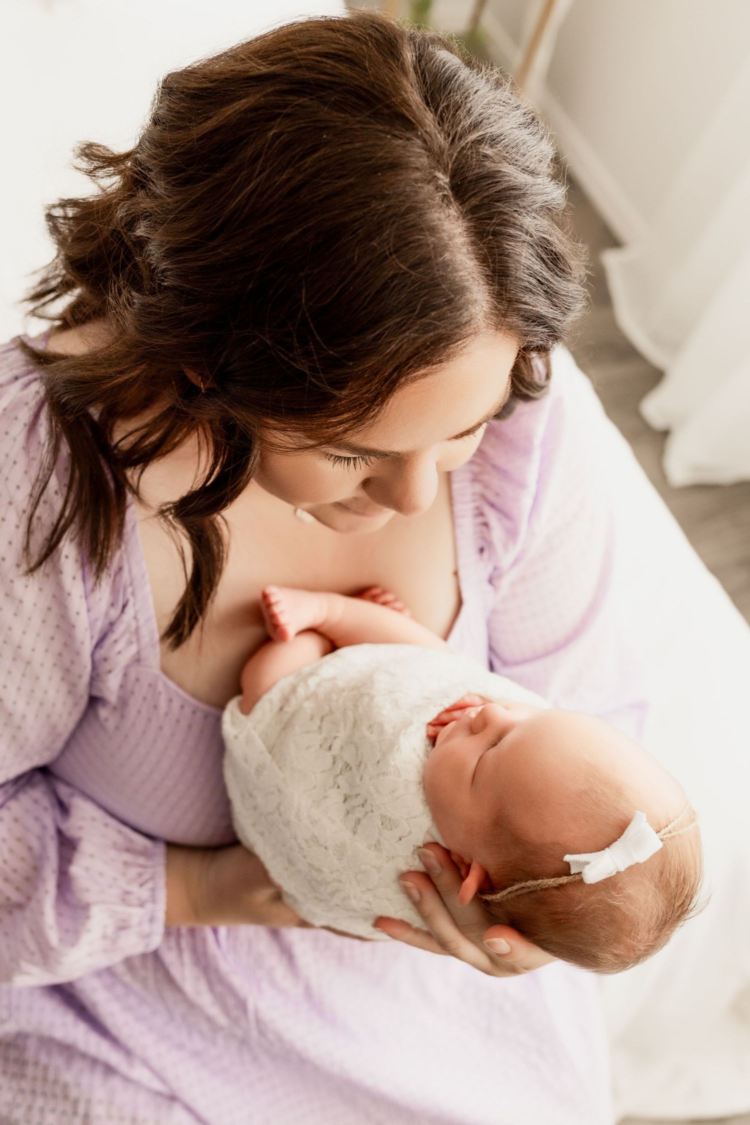 A mother looks down at her newborn daughter, swaddled in a white wrap with a cute bow headband during newborn photo session.
