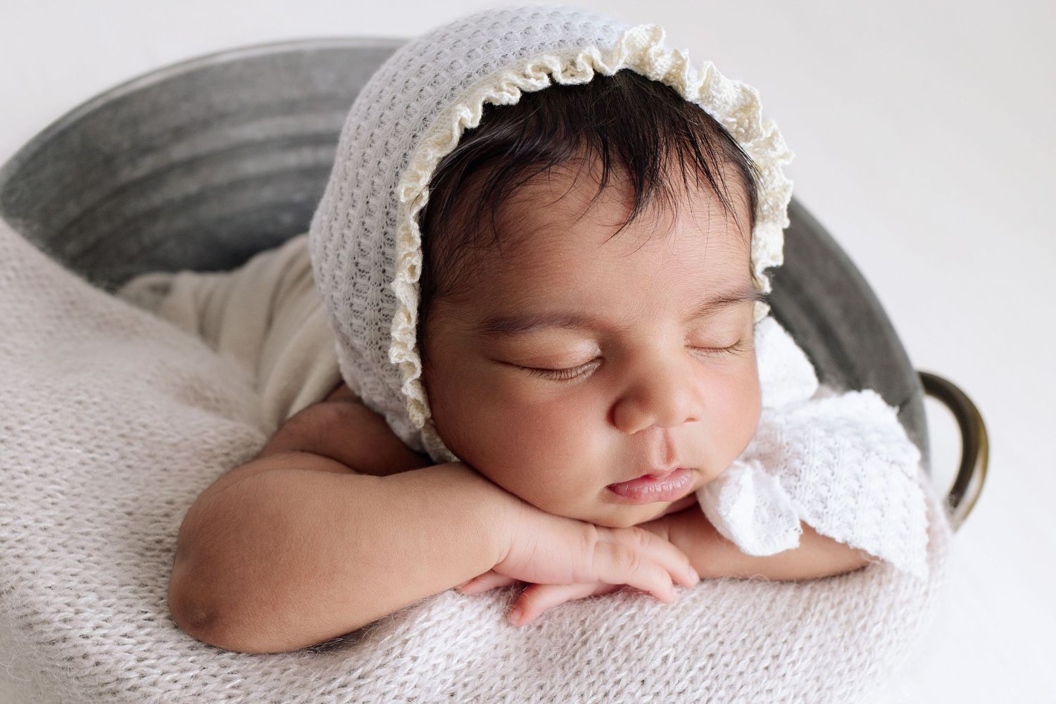 Sleeping baby girl with old-fashioned lace edged bonnet rests her hands on edge of the metal bucket she is in, head resting on her hands.