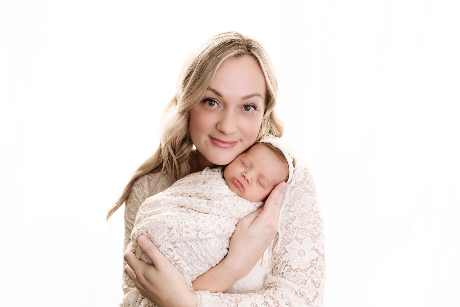 Luxury newborn photo session of baby girl swaddled in a cream lace wrap. Mom holds baby's face cheek to cheek. Bright white background.
