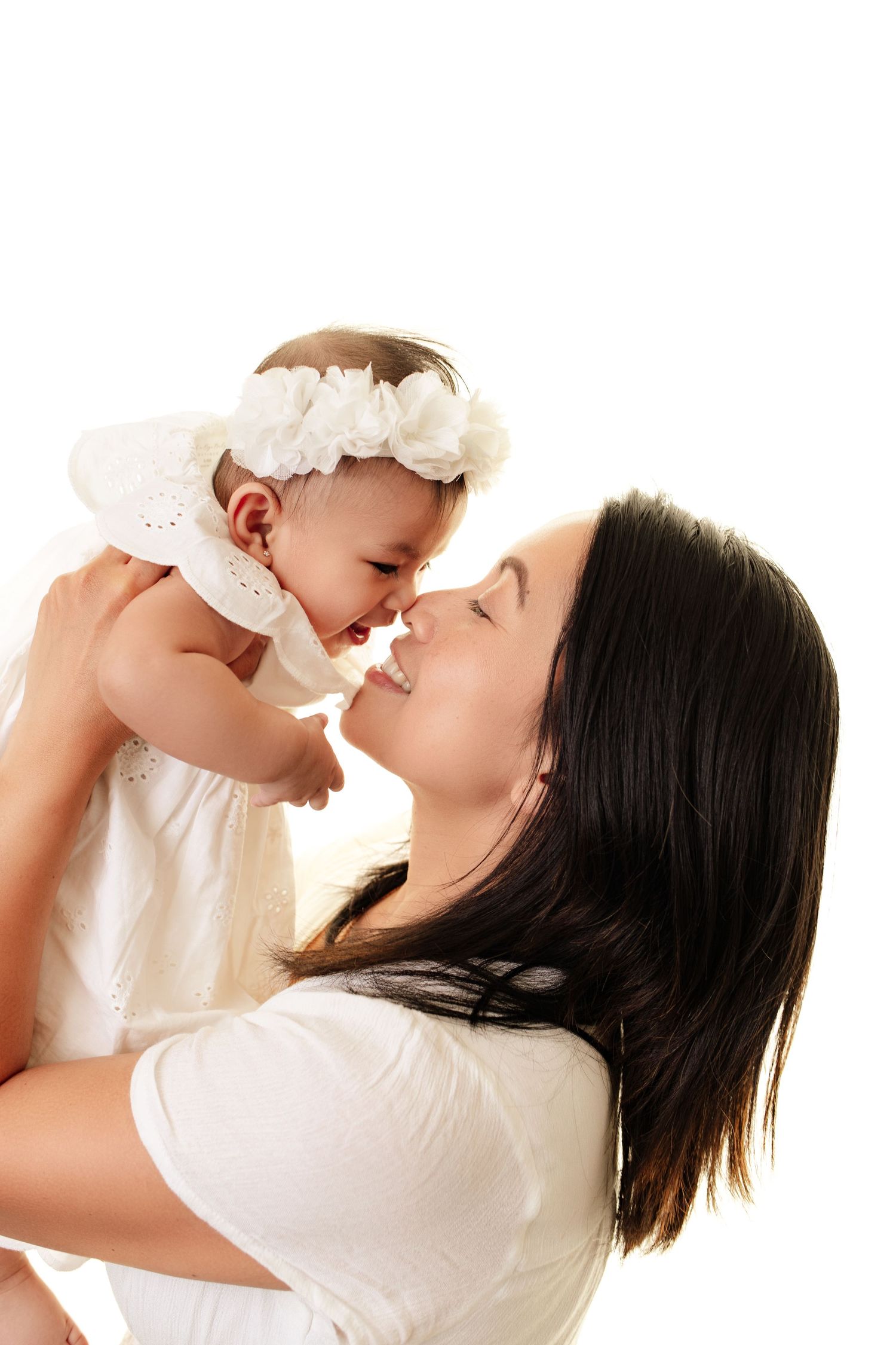 Beautiful mom and baby photo, mom is holding baby up, and they are nose to nose with a bright white background. Both are smiling.