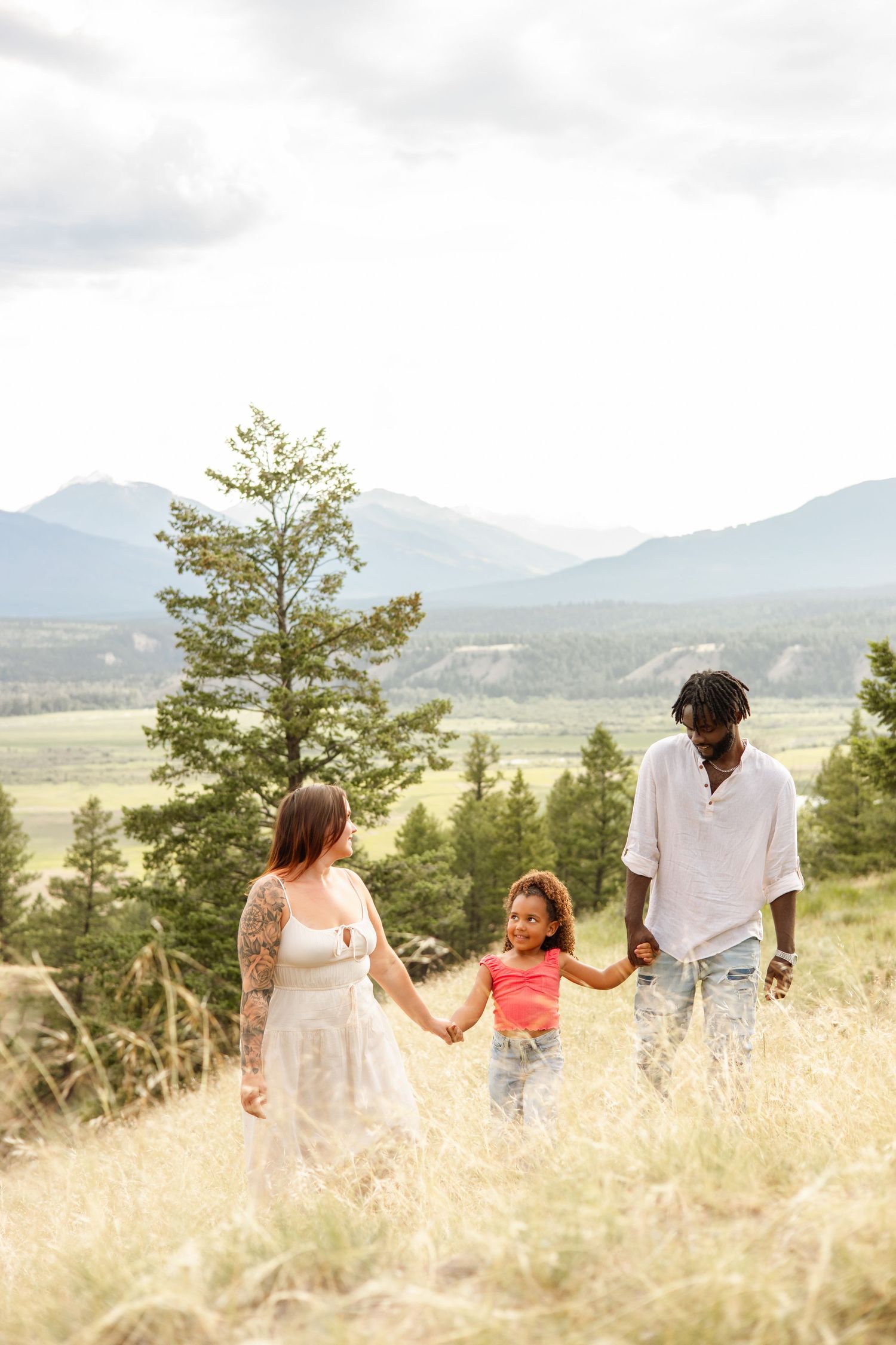 Gorgeous multiracial family photography of mom, dad and daughter holding hands and walking on hillside with beautiful mountain backdrop.