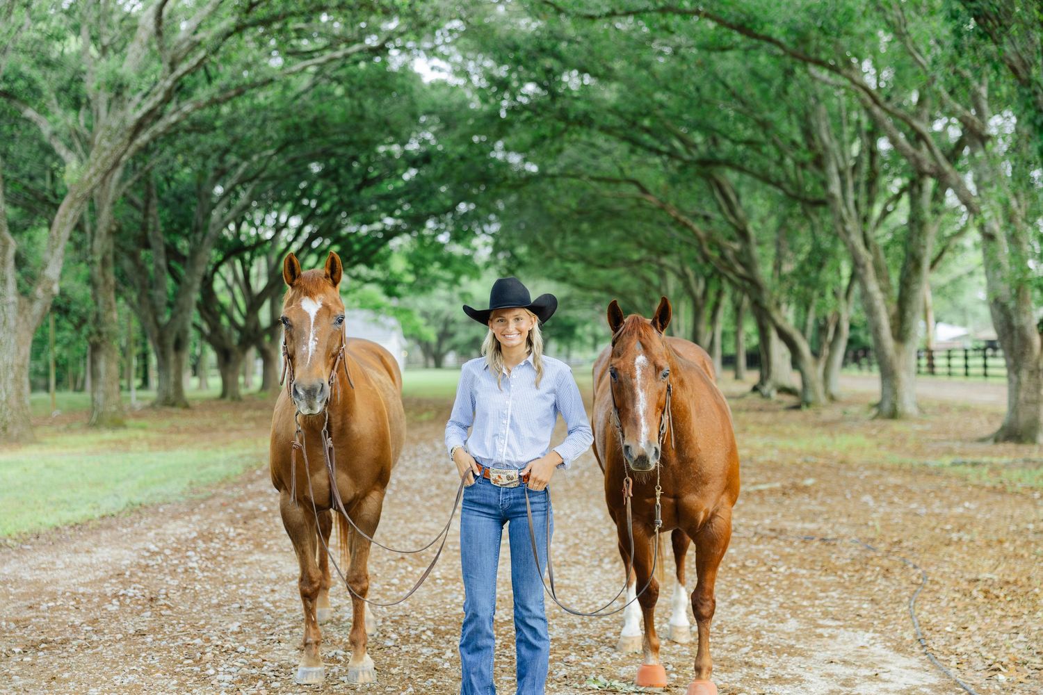 Young woman in cowboy hat leads two chestnut horses down a tree-lined path on a sunny day.