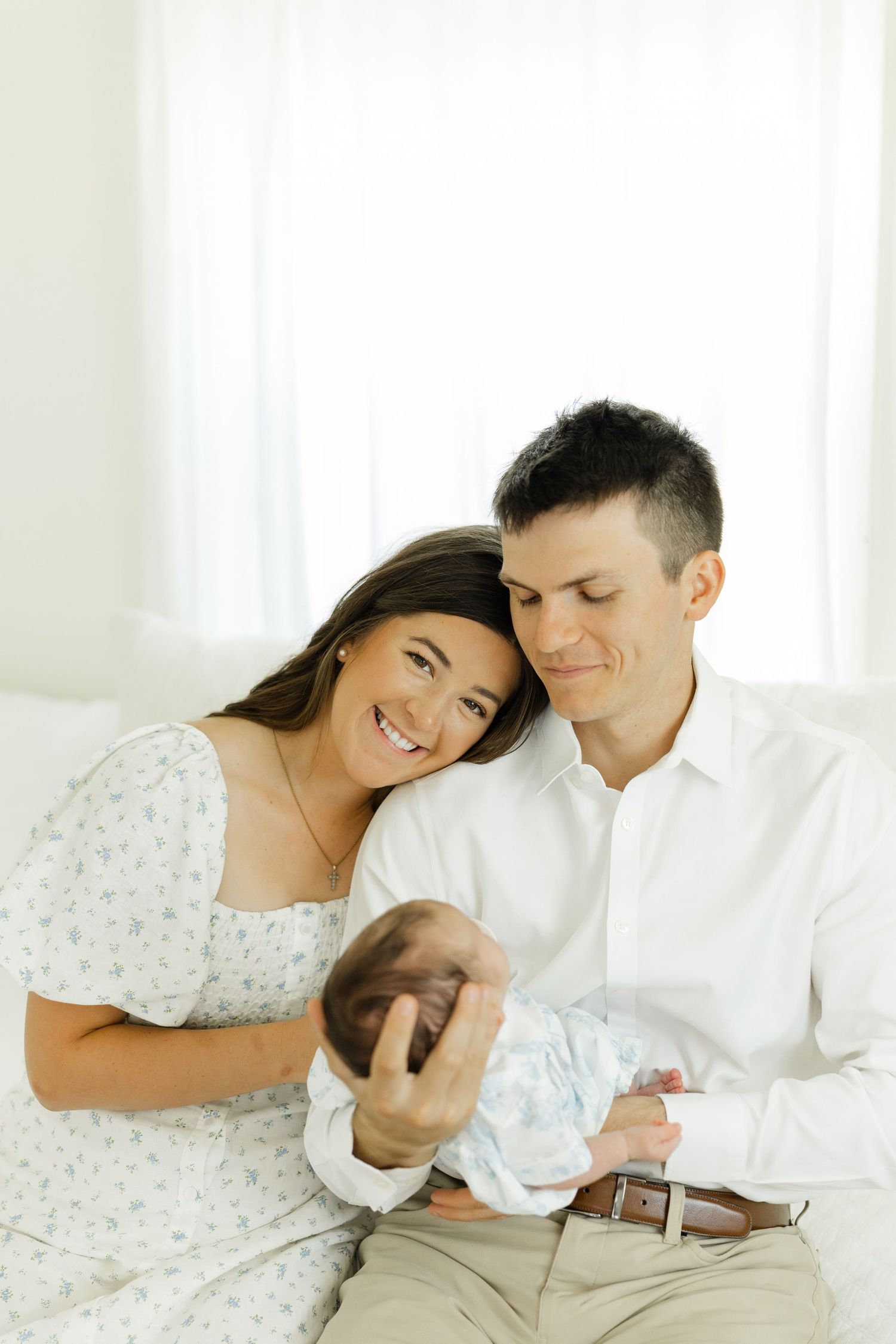 Smiling new parents sit together on a bed, lovingly gazing at their swaddled newborn baby.