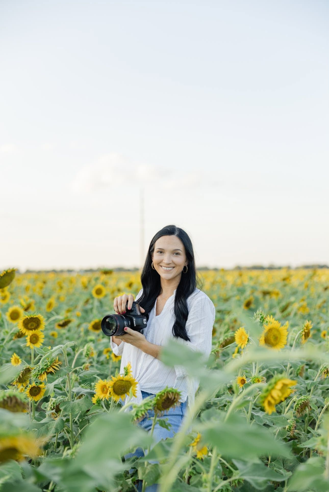 Smiling female photographer holding camera in a vibrant sunflower field at dusk.