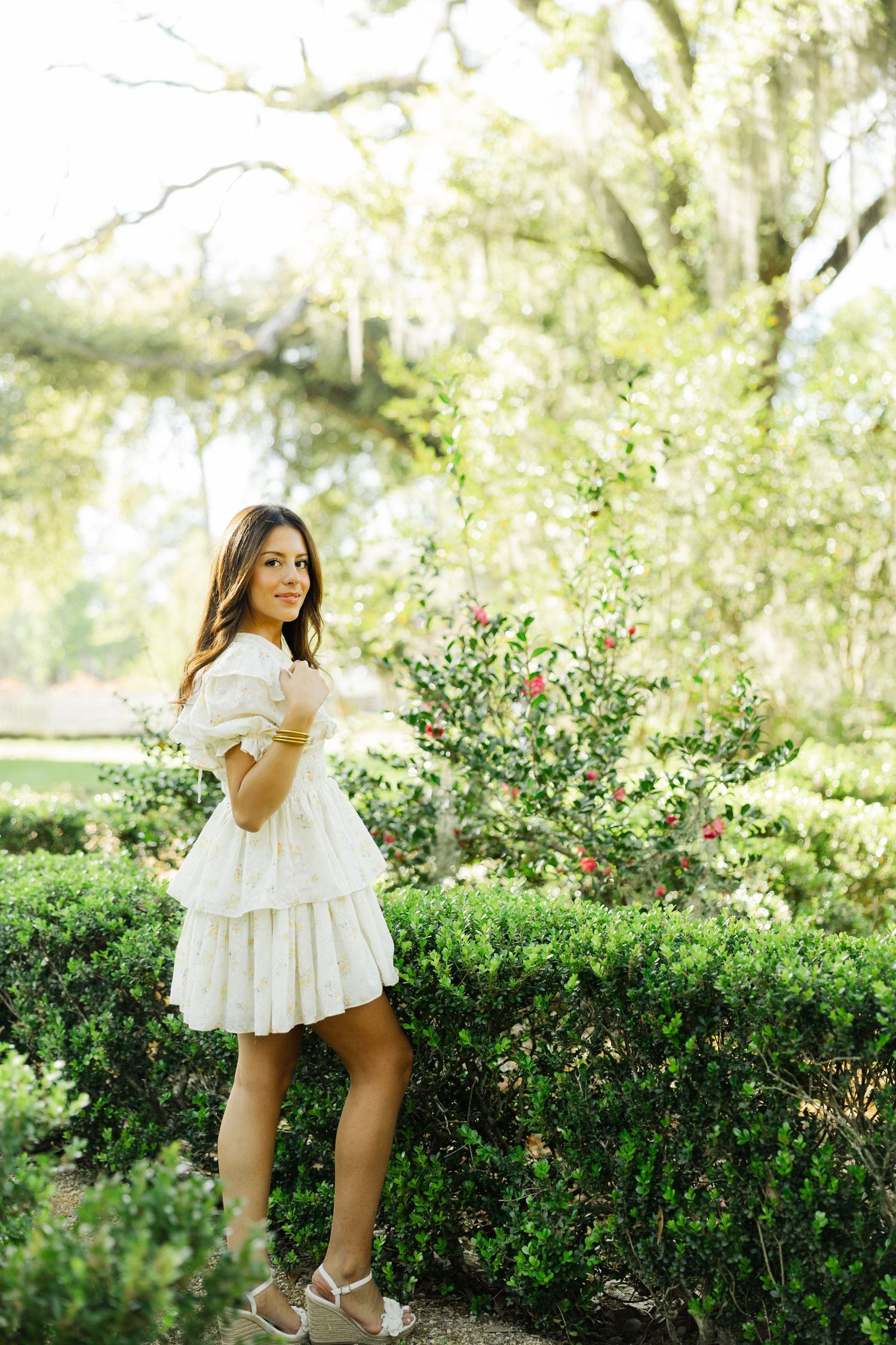 Young woman in white tiered mini dress posing beside manicured hedges in a sunlit garden.