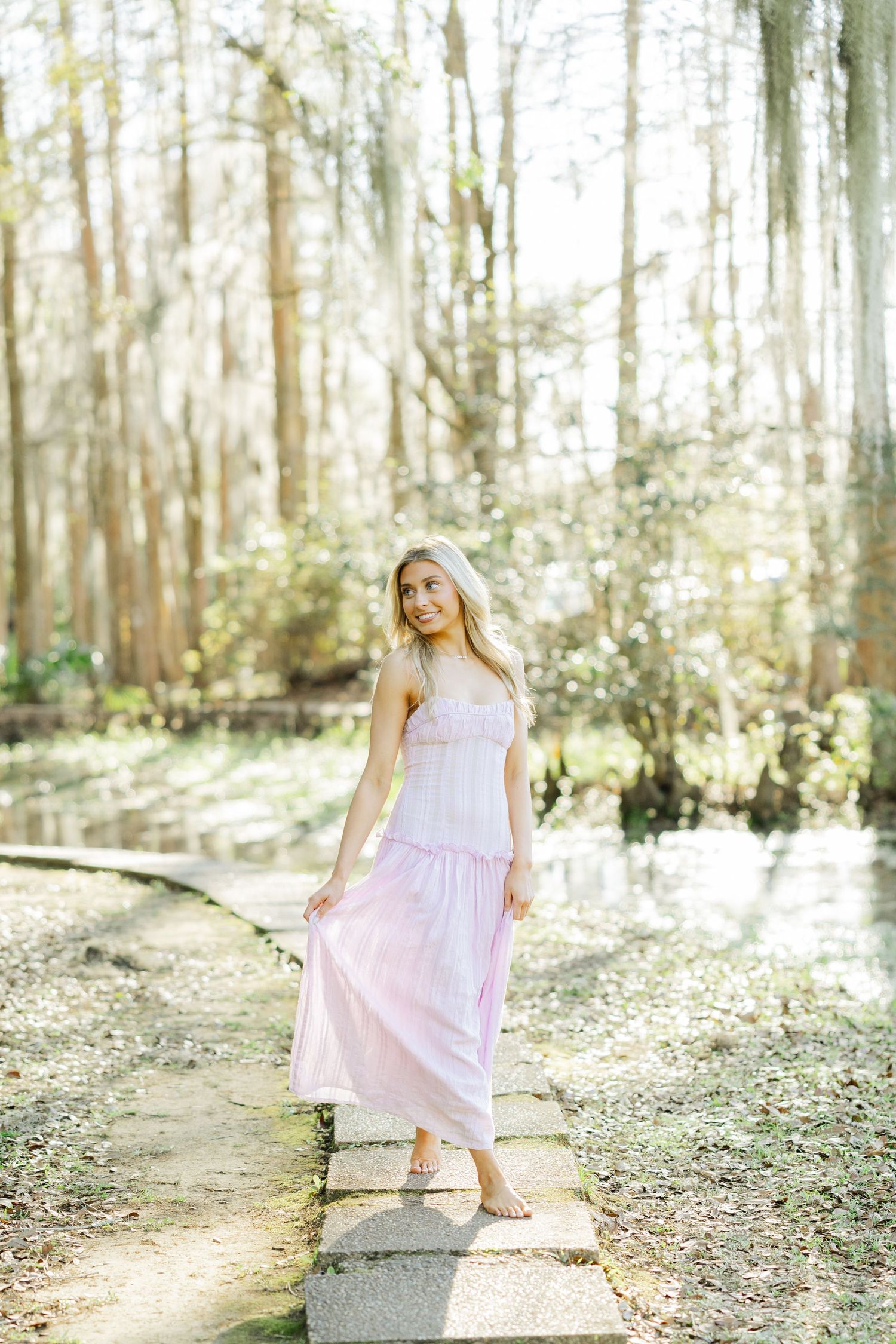 Blonde woman in a flowing pink maxi dress standing barefoot on stone path in a sunlit forest.