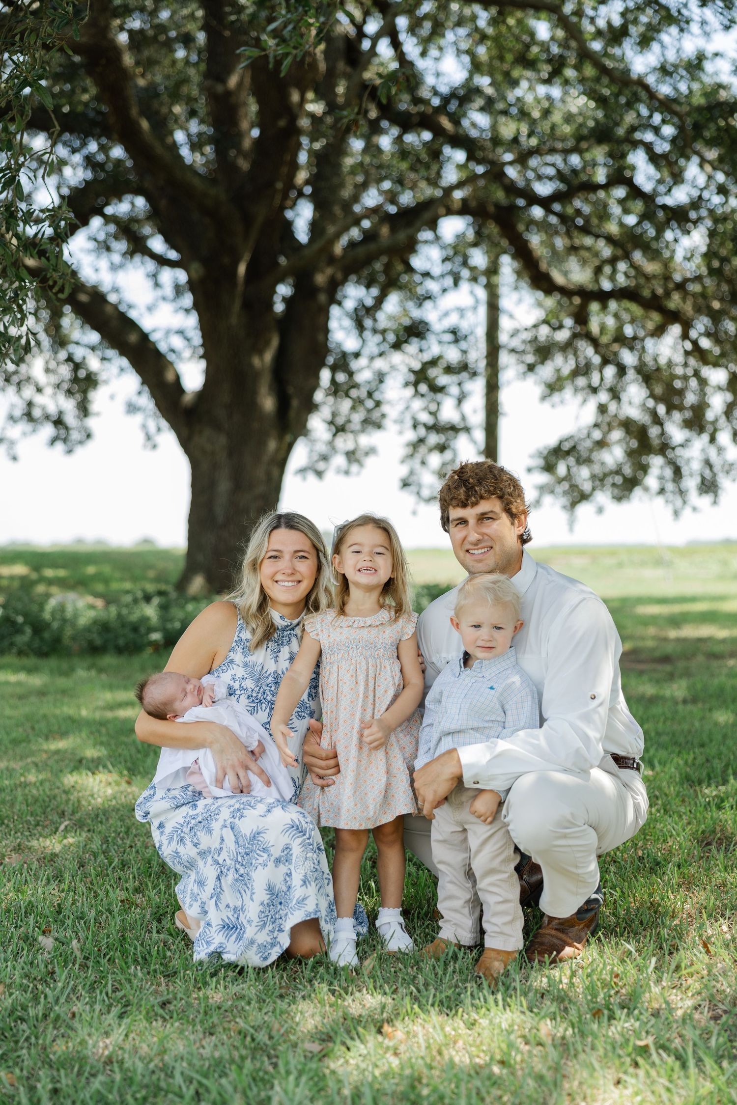 A happy family of five poses under a large oak tree, including a newborn baby and two young children.