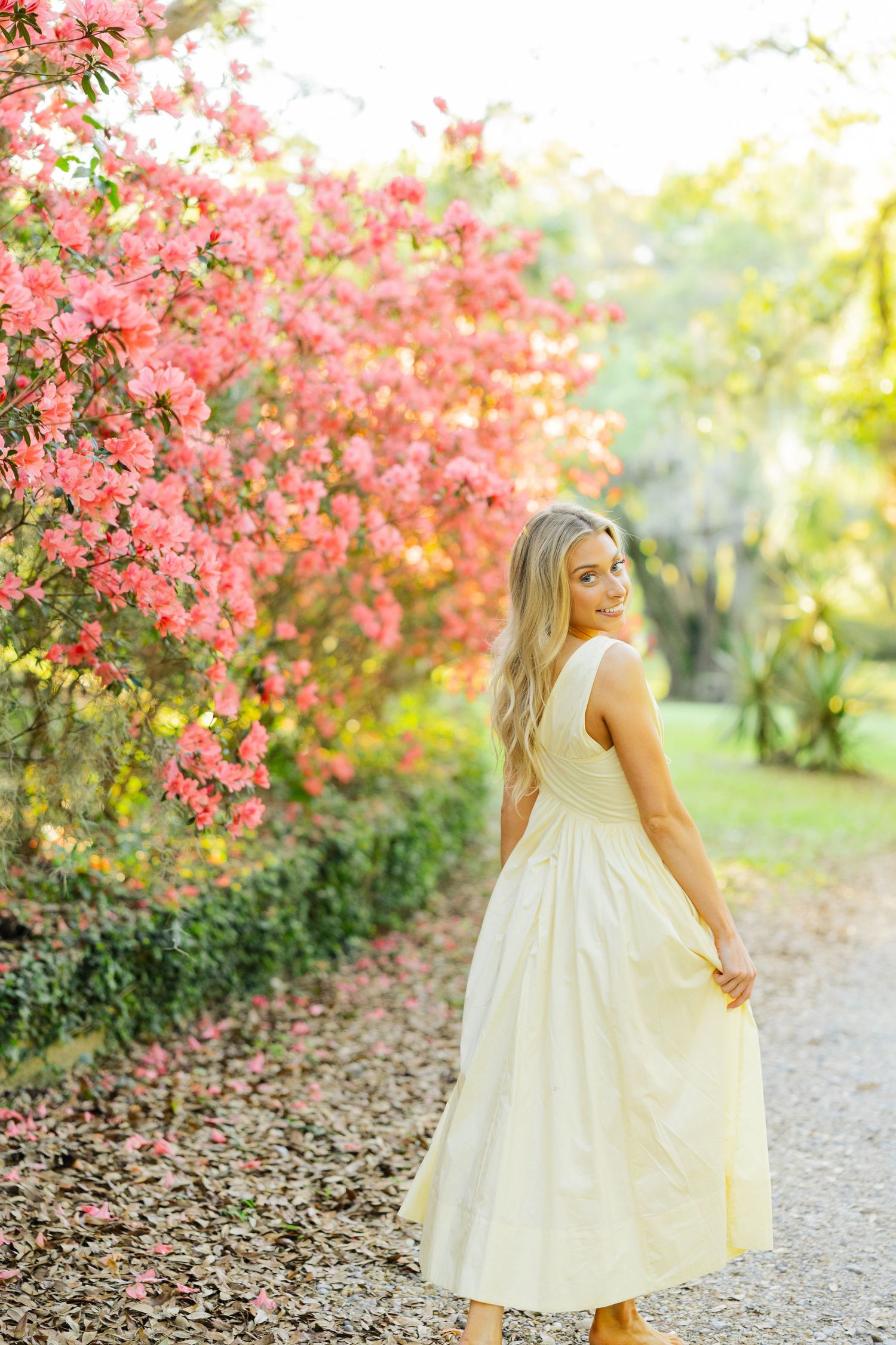 Smiling blonde woman in yellow dress twirls near blooming pink azaleas on a garden path.