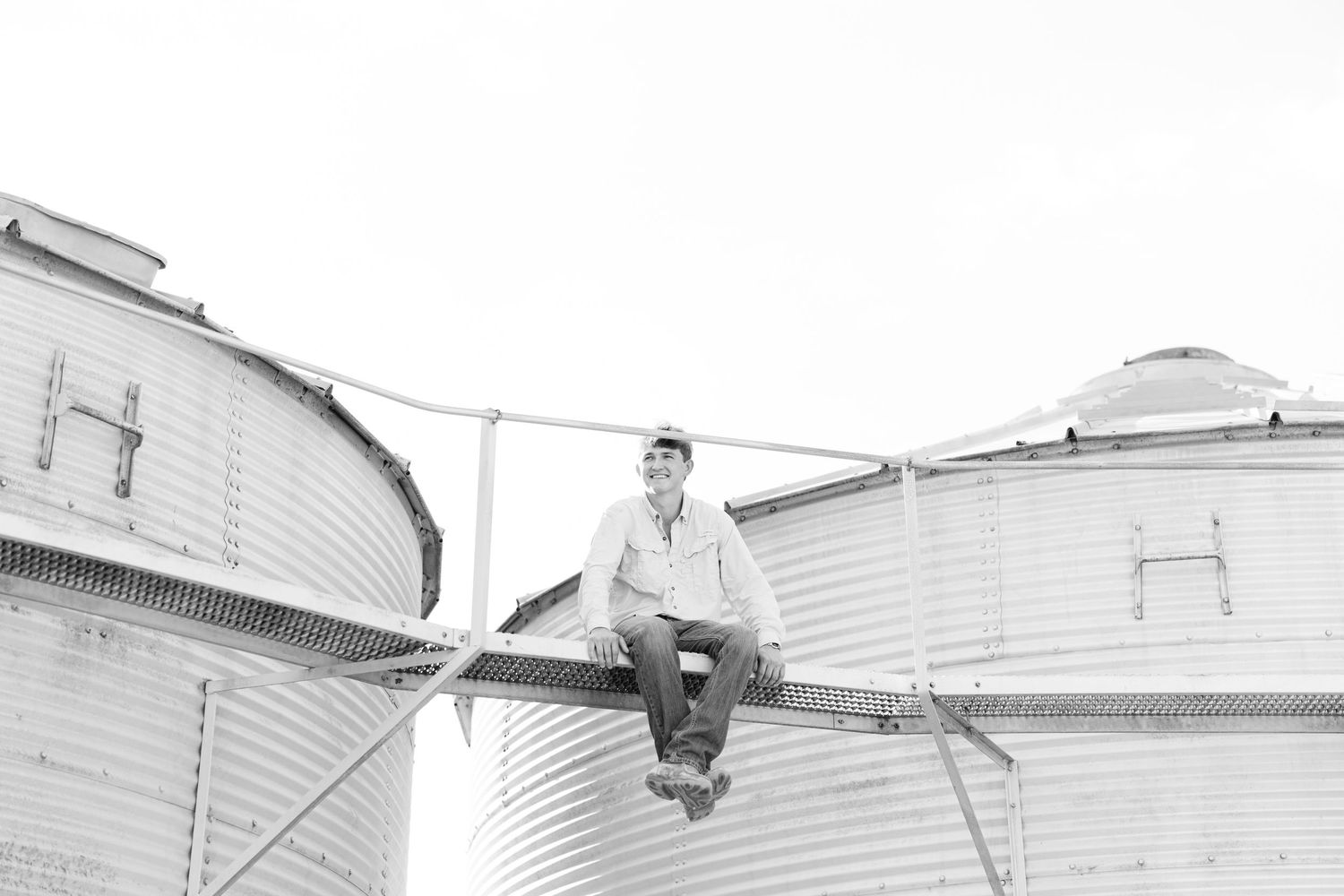Young man sits atop grain silos in black and white rural farm photography.