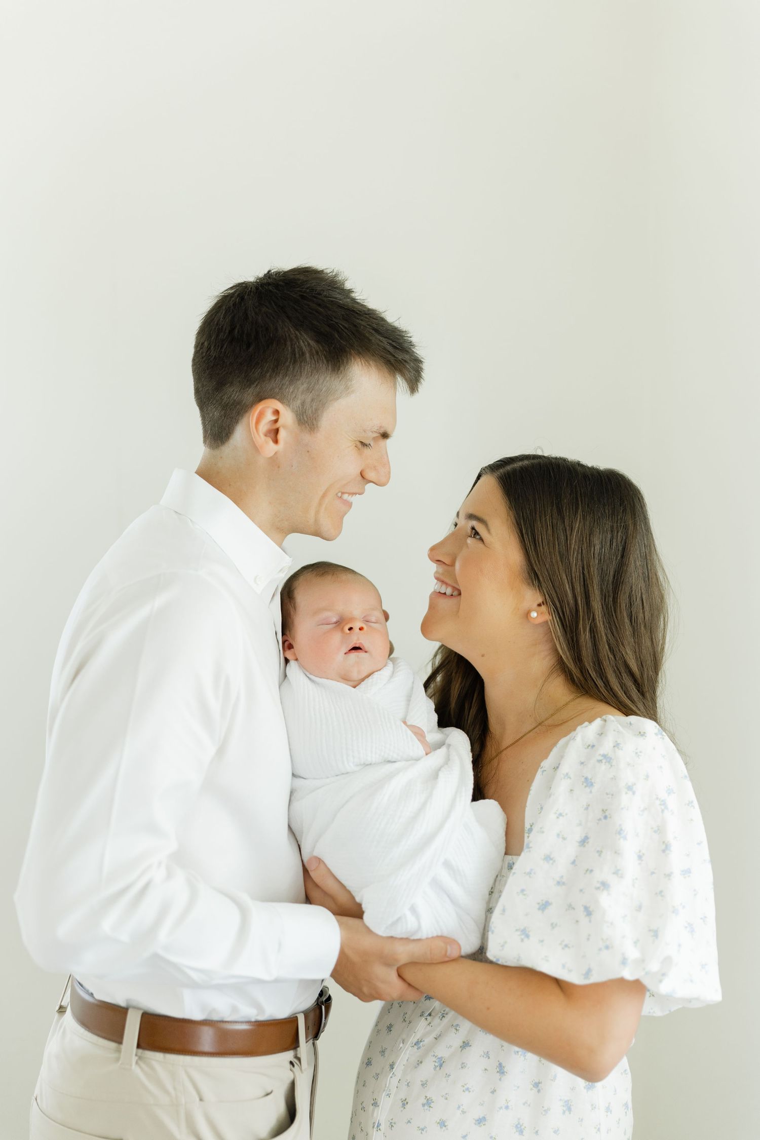 Happy young couple in white outfits smile at each other while holding their swaddled newborn baby.