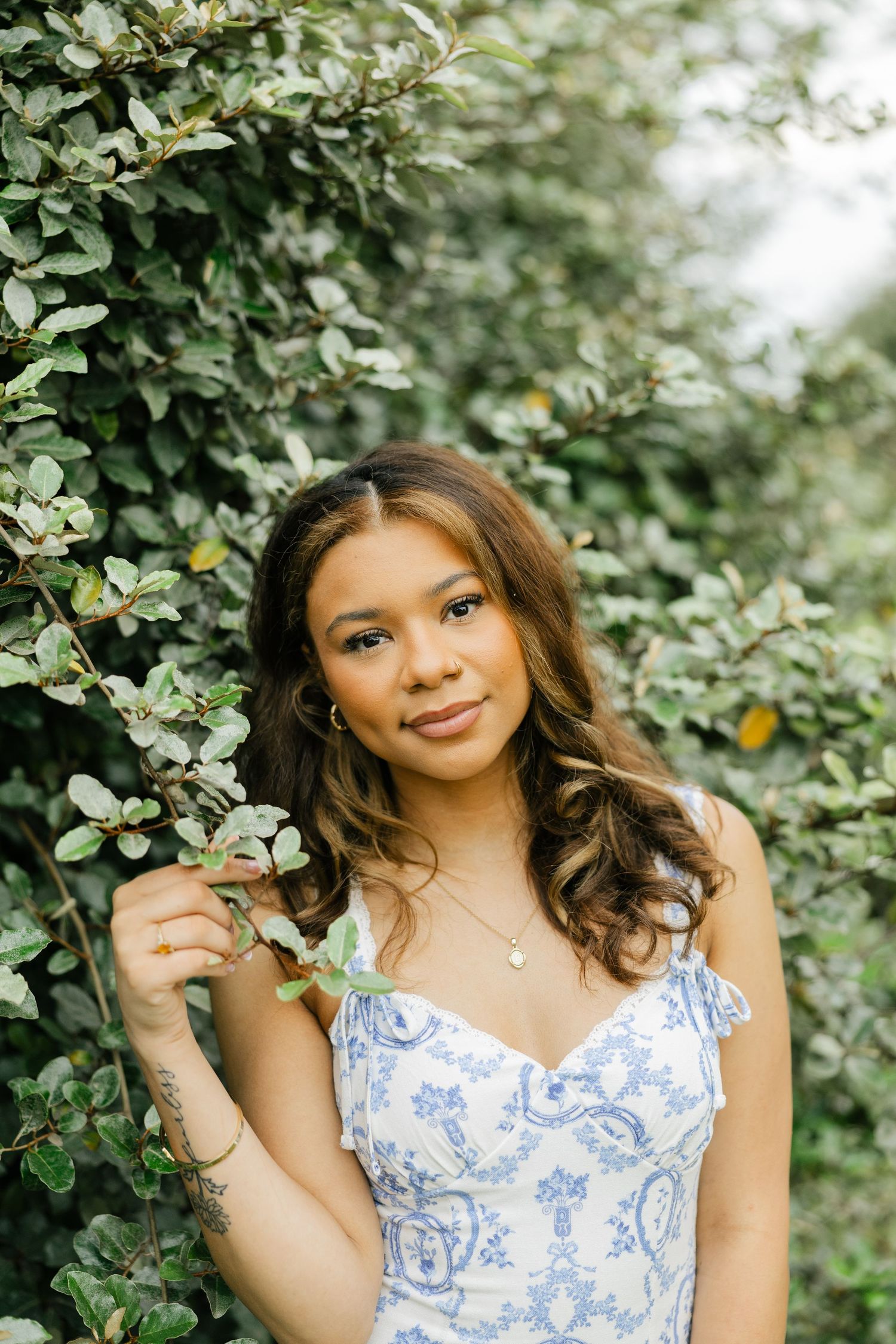 Young woman in blue floral sundress posing beside lush green leafy bushes outdoors.