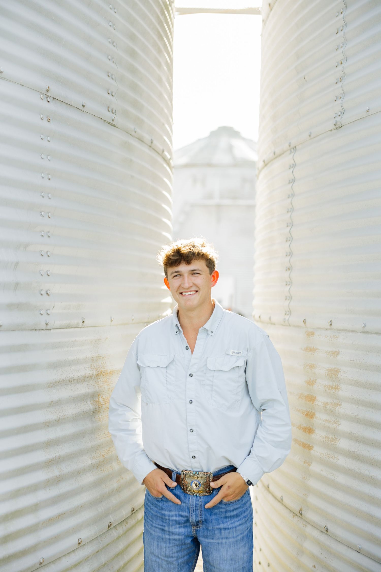 Smiling young man in a western shirt and jeans standing between two large metal grain silos.