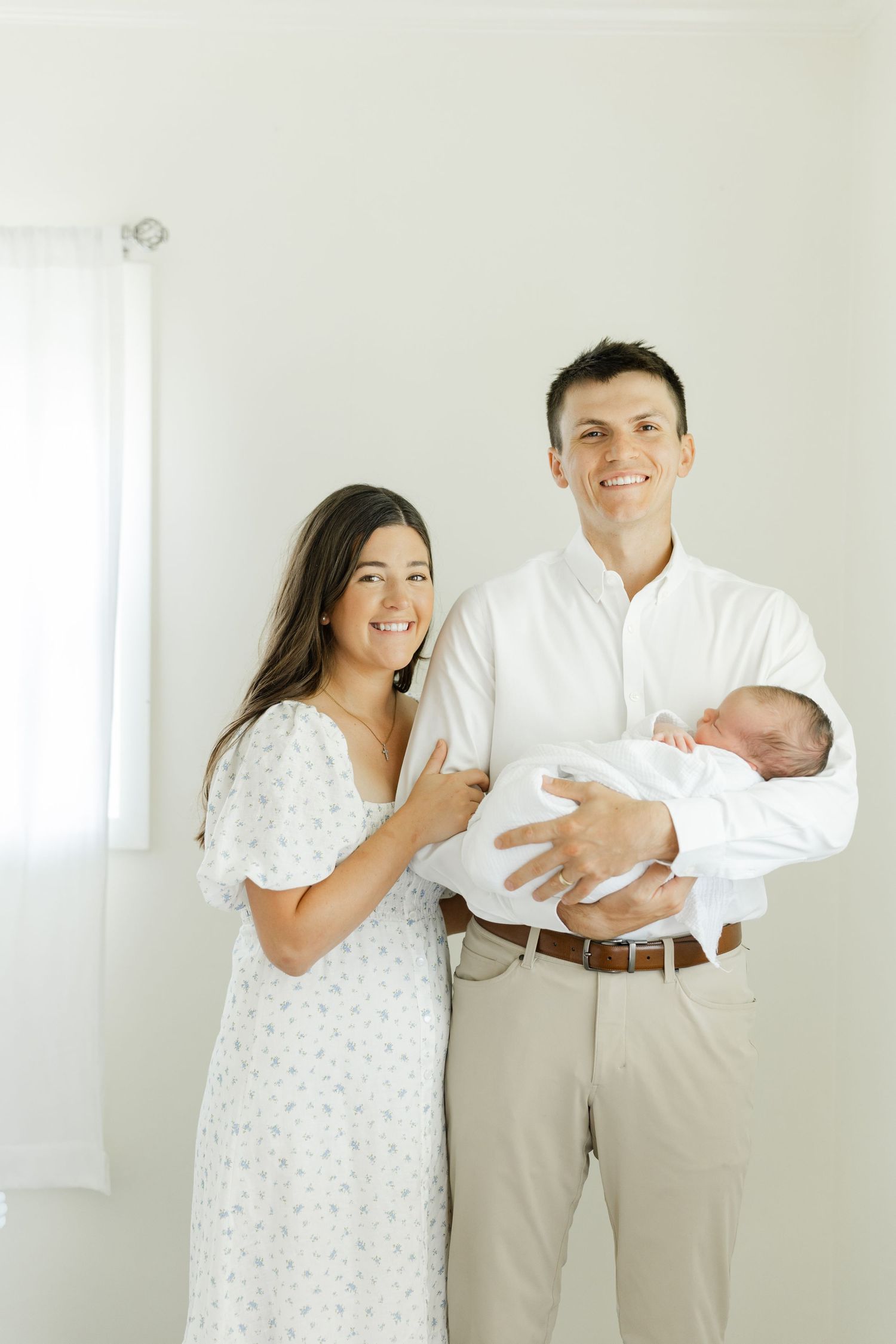 Smiling new parents pose indoors, father holding swaddled newborn while mother in floral dress stands beside him.