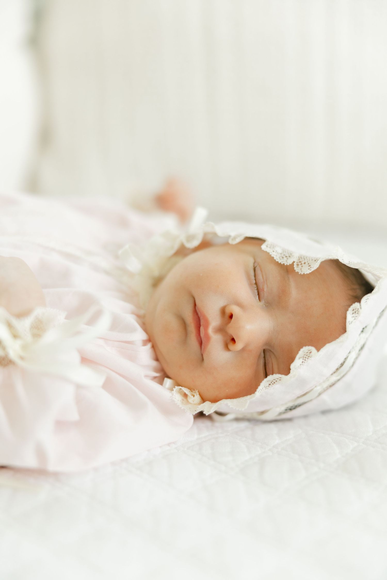 Sleeping newborn baby in pink gown and white lace bonnet lying on white quilted blanket.